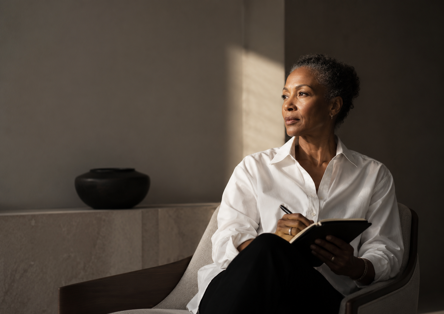 An older woman with gray curly hair, wearing a white button-up shirt, sitting in a modern room, looking thoughtfully out of the window while holding a notebook and pen.