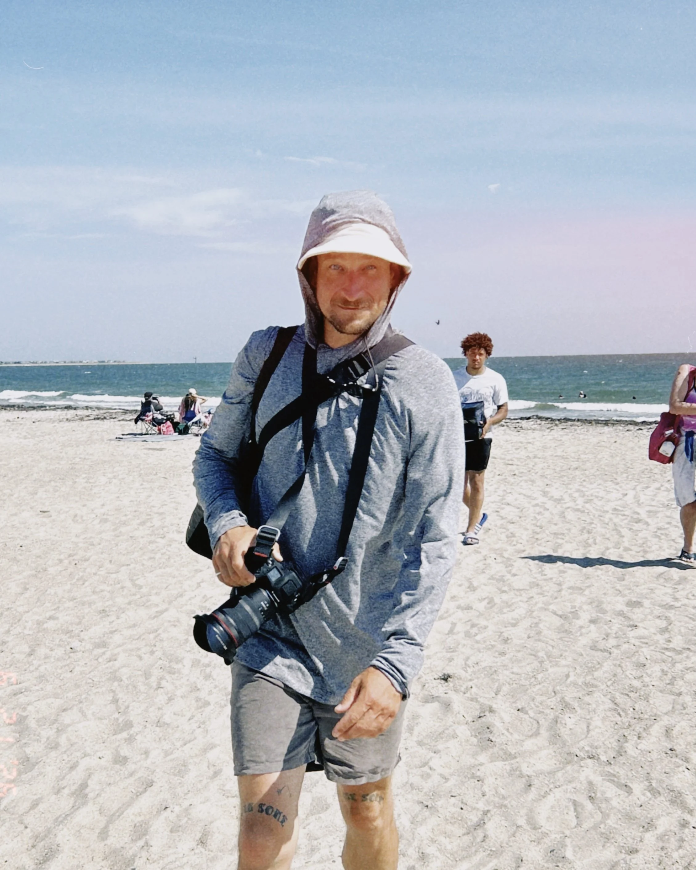 A man wearing a gray hooded jacket and shorts, holding a camera with a large lens, stands on a beach with the ocean in the background. Other people are visible in the background.