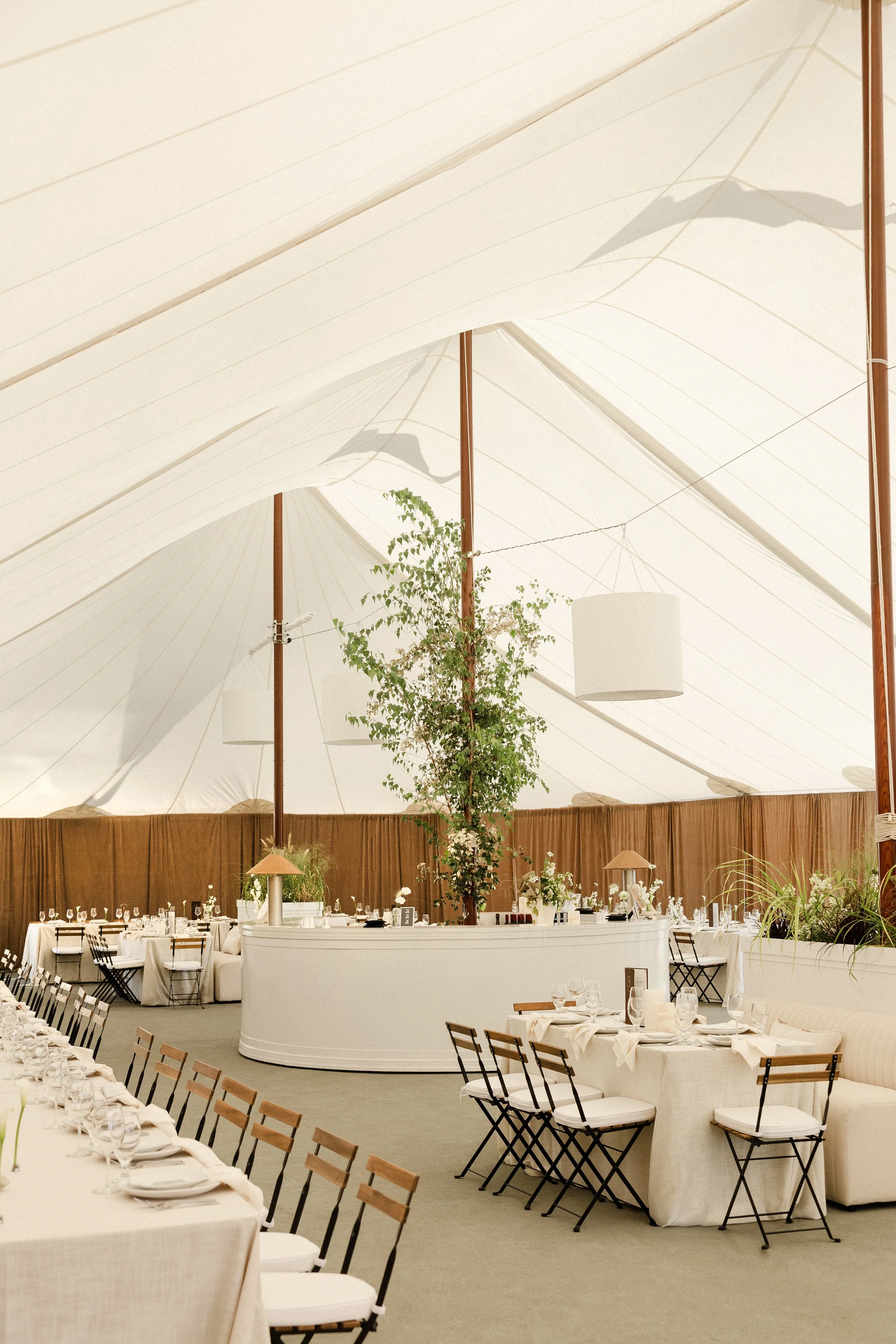 Elegant dining area inside a white tent, featuring a tall central plant, intricate lighting, and tables set with white linens, glassware, and dinnerware.