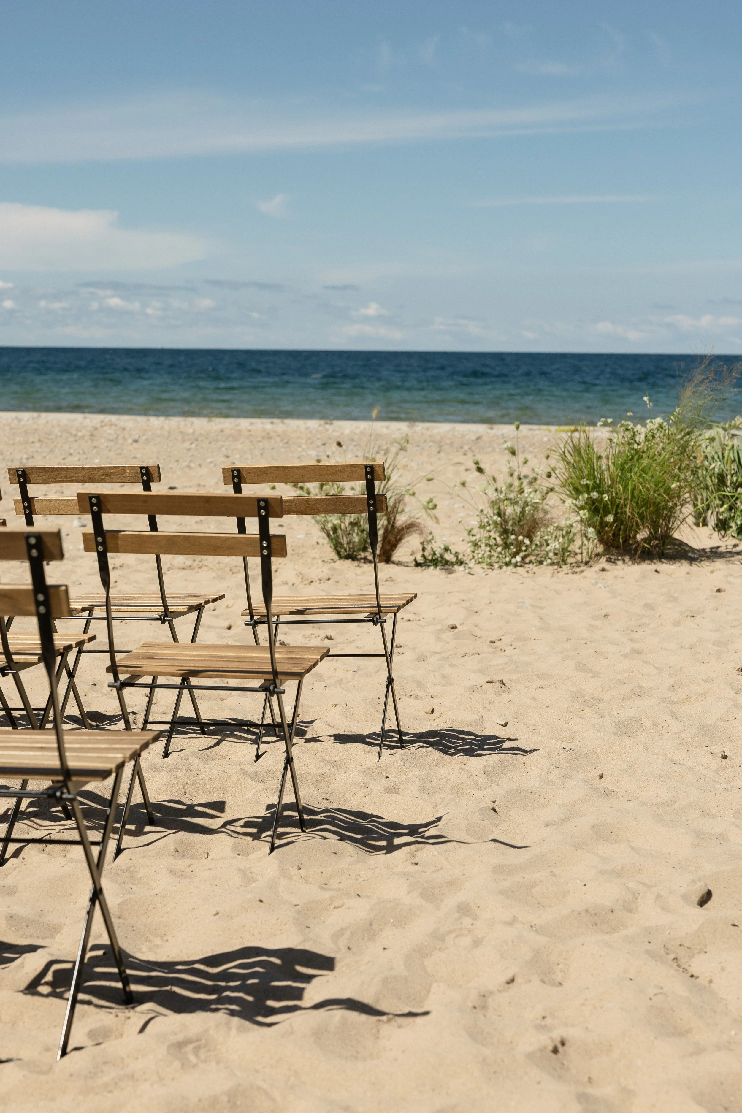 Empty wooden chairs on sandy beach facing the ocean with some green plants near the shoreline under a partly cloudy sky.