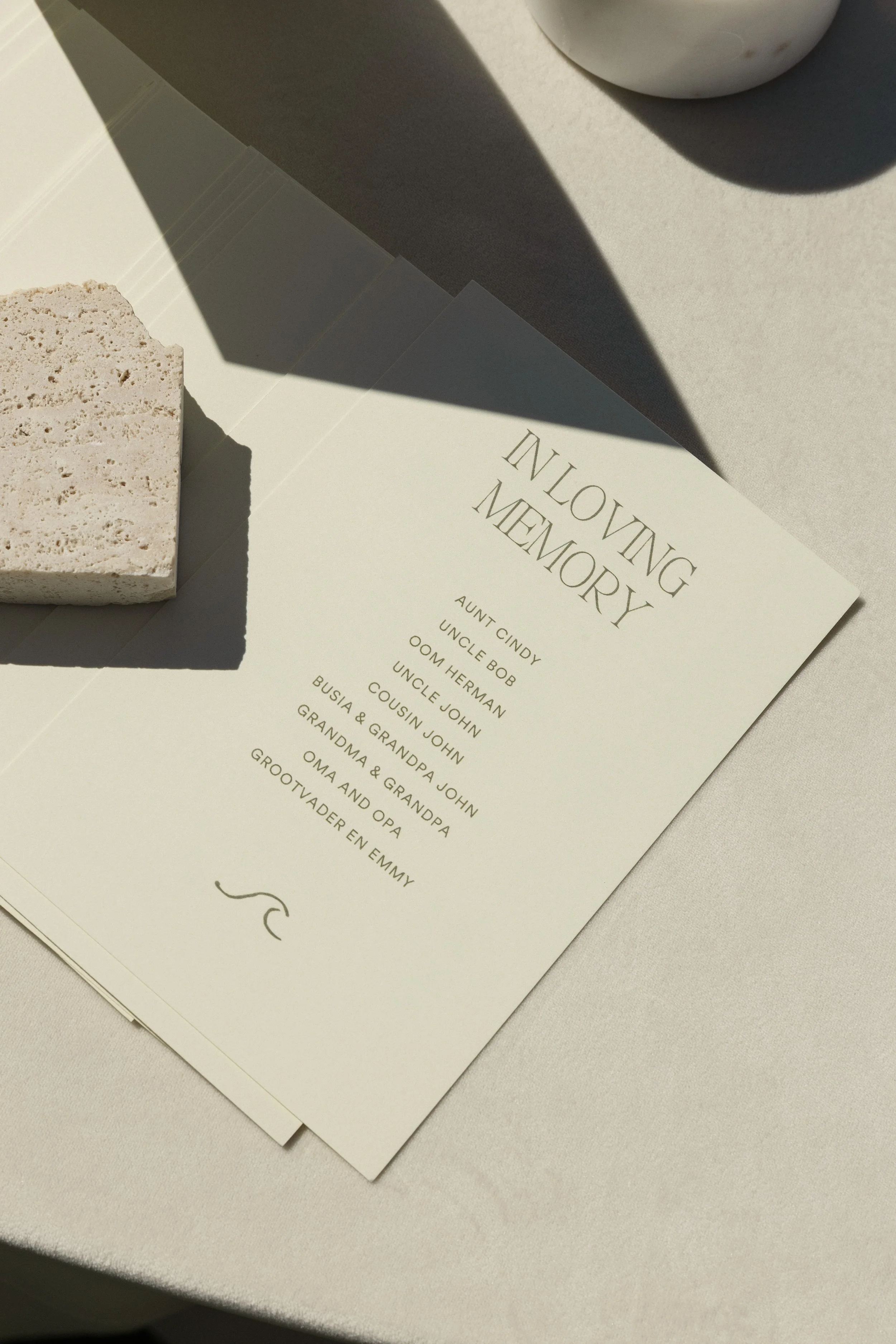 A memorial program titled 'Including Memory' on a white table, accompanied by a small beige stone block. The program lists names of family members, including Aunt Cindy, Uncle Bob, Uncle Herman, Uncle John, Cousin John, Busia & Grandpa, Grandma & Grandpa, Oma and Opa, and Grootvader en Emmy.