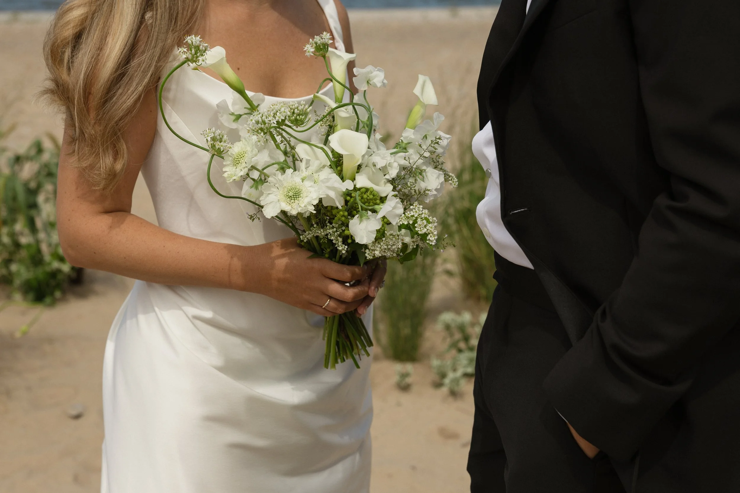 A bride holding a bouquet of white flowers, standing next to a man dressed in a black tuxedo, with a beach in the background.