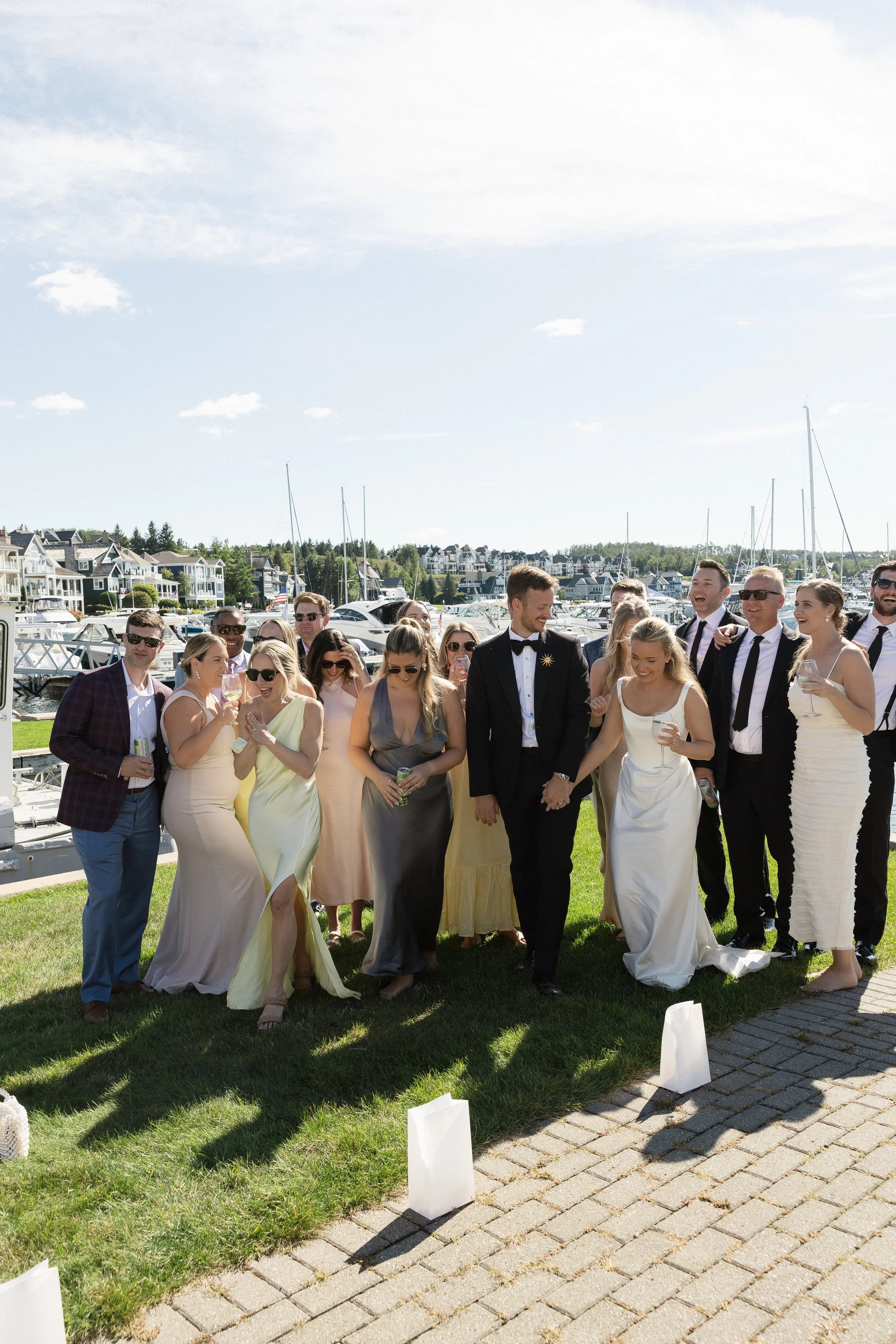 Group of people dressed in formal attire, some wearing sunglasses, standing on grass with a marina and boats in the background, celebrating outdoors on a sunny day.