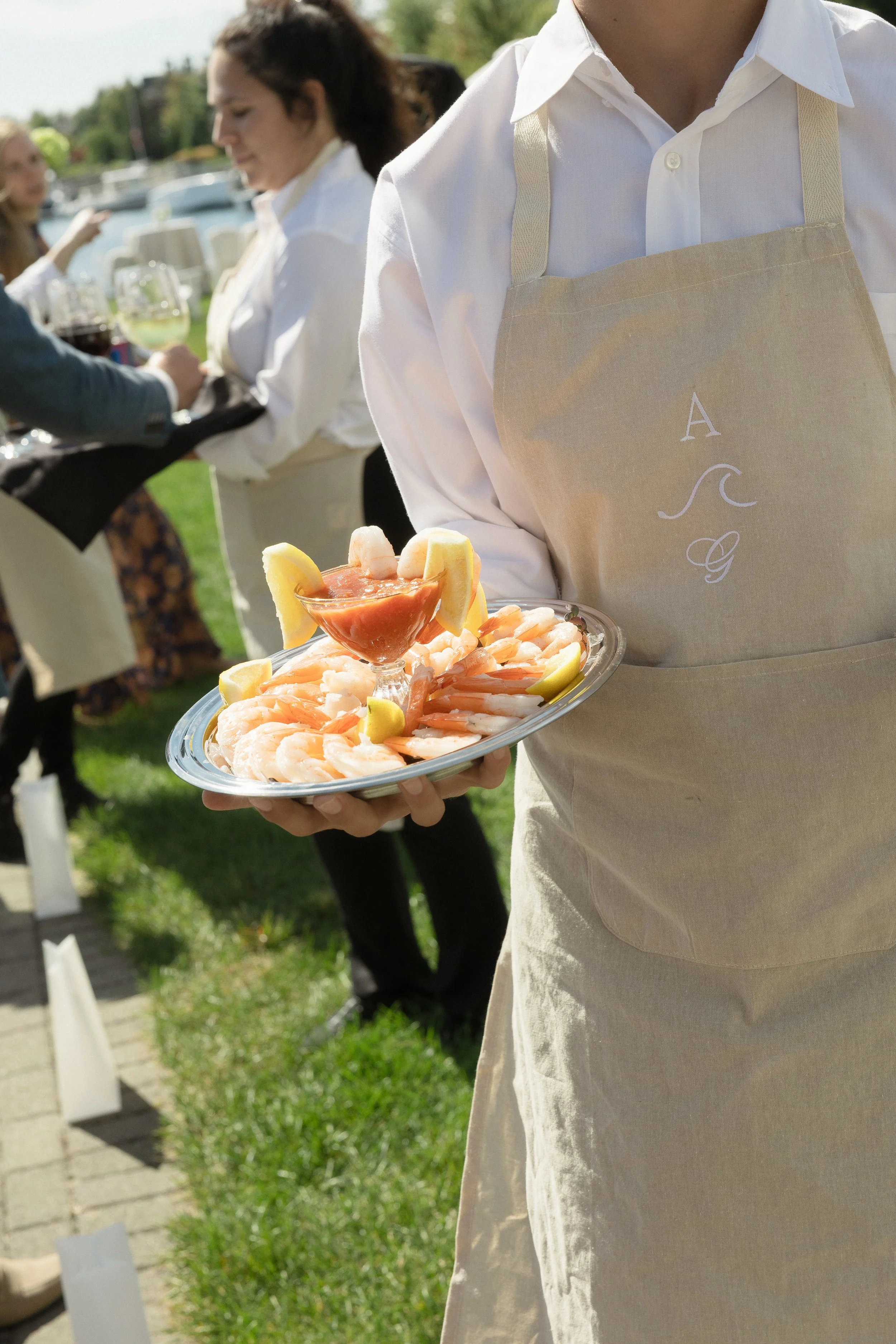 A waiter wearing a beige apron with an embroidered monogram offers a tray of shrimp garnished with lemon wedges, with a small bowl of cocktail sauce in the center, at an outdoor gathering by the water.