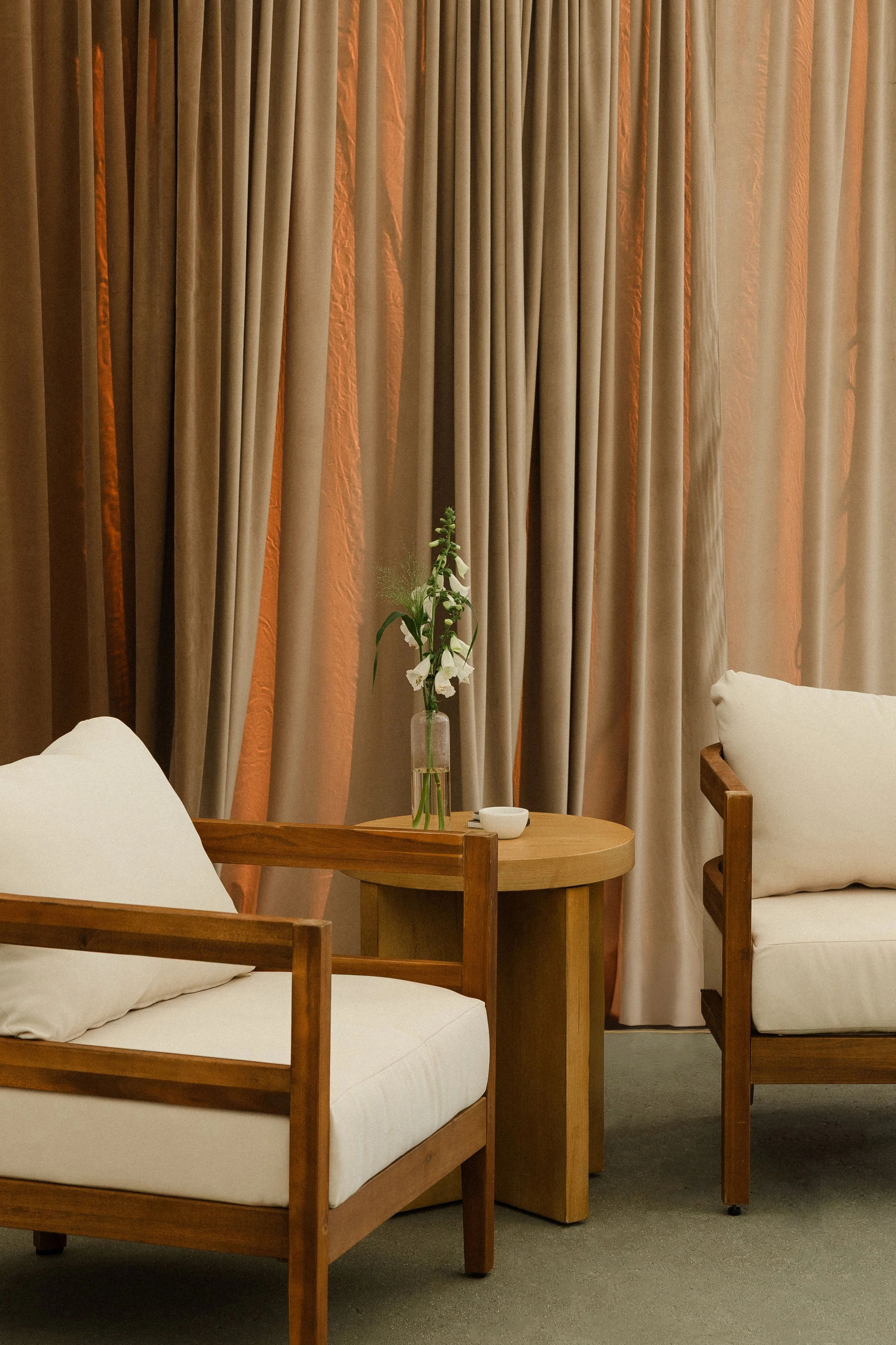 Living room corner with two wooden armchairs with white cushions, a small round wooden side table with a vase of white flowers and a small white dish, and beige curtains hanging in the background.