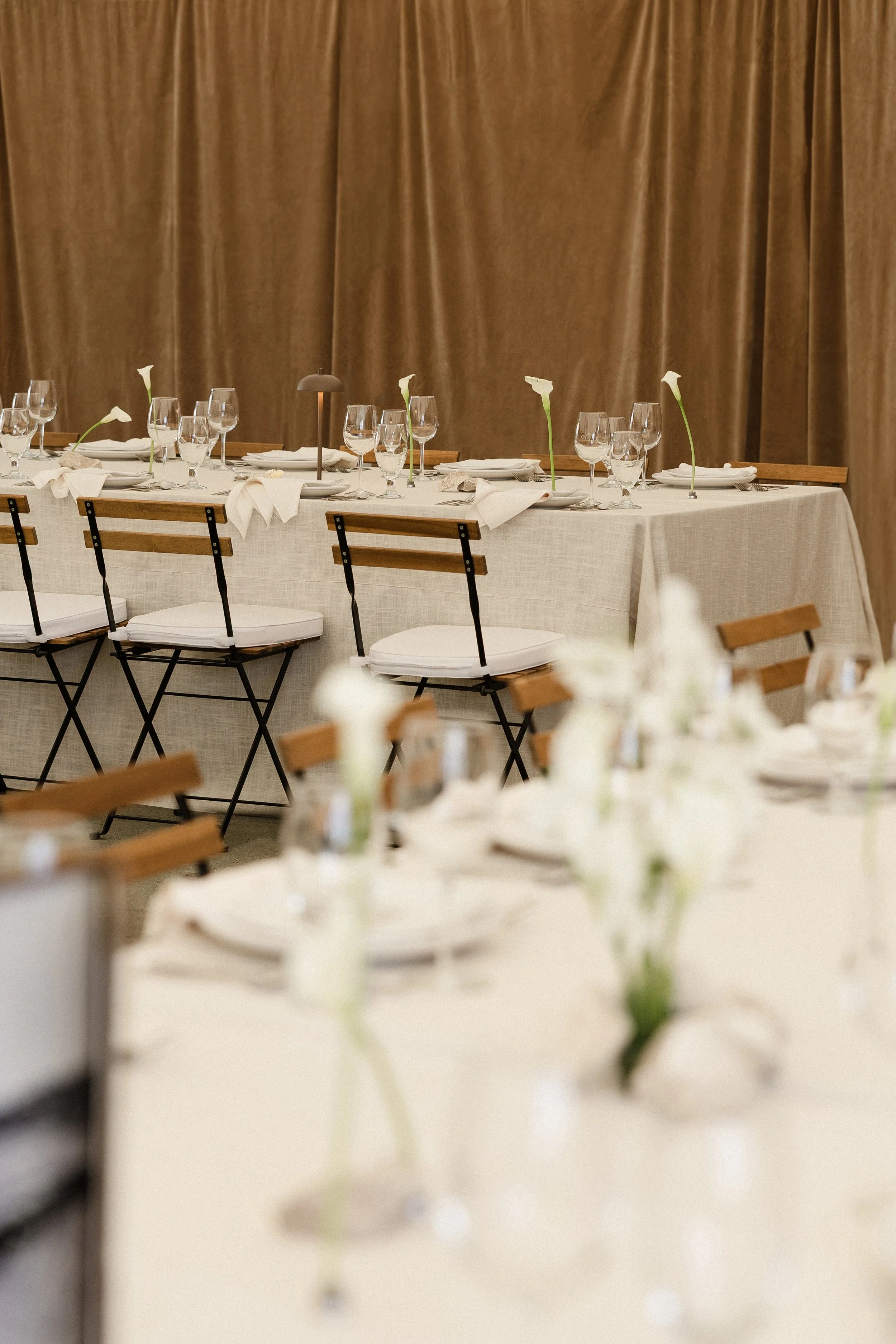 Elegant dining table set for an event, decorated with white flowers and wine glasses, with a beige curtain backdrop.