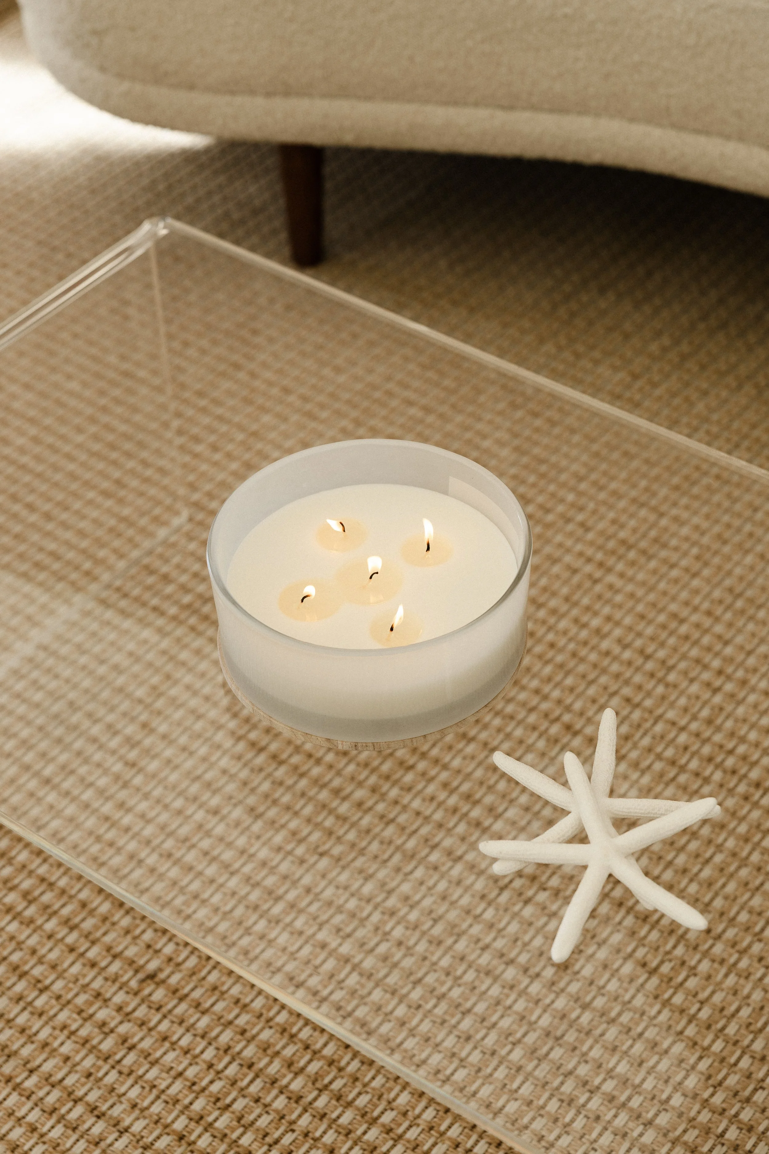 A white candle with five lit wicks in a transparent glass holder, placed on a glass side table with a starfish next to it, on a beige textured surface.