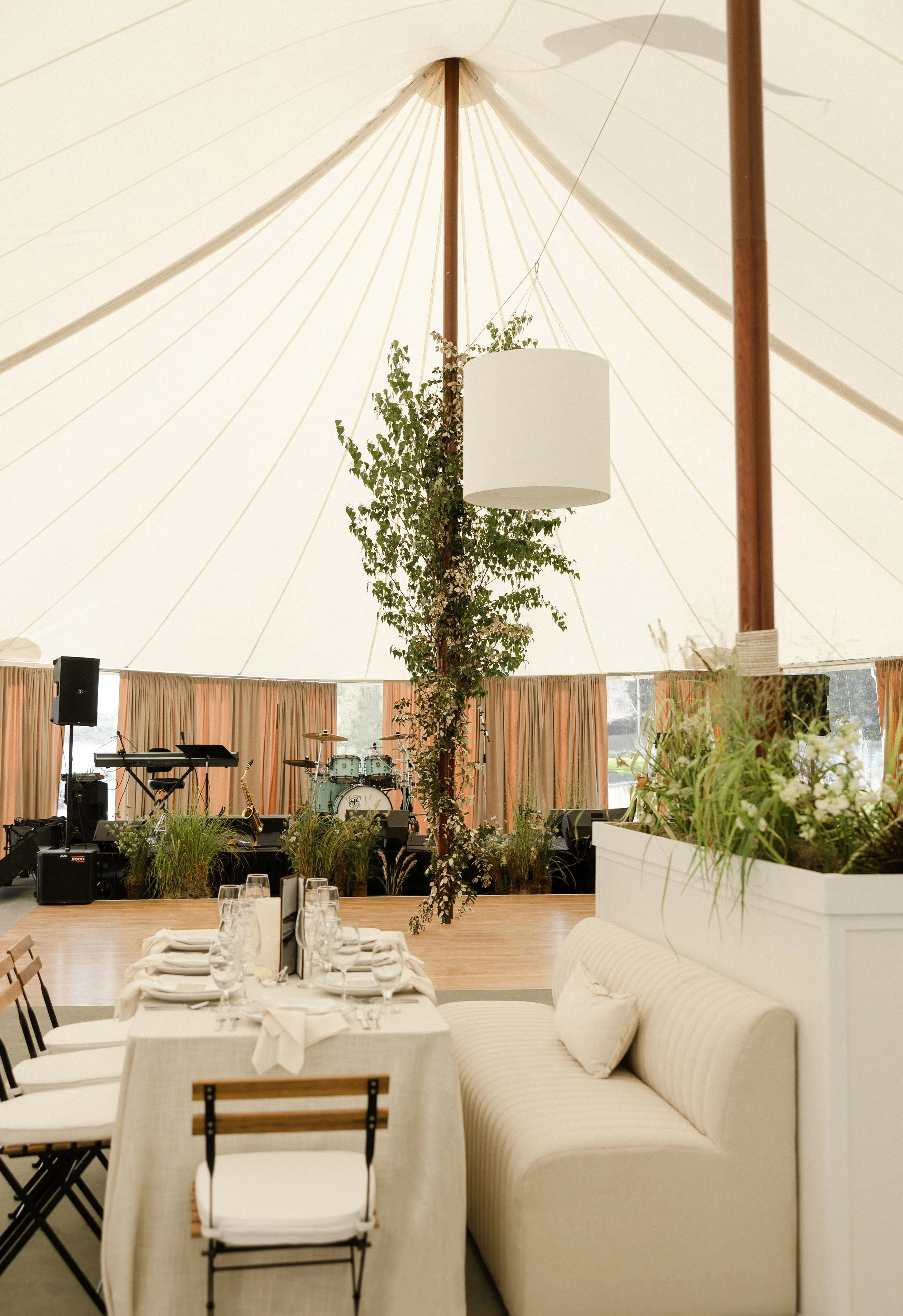Interior of a tent decorated for an event, with a stage for live music, a dining table set with glassware, and greenery accents.