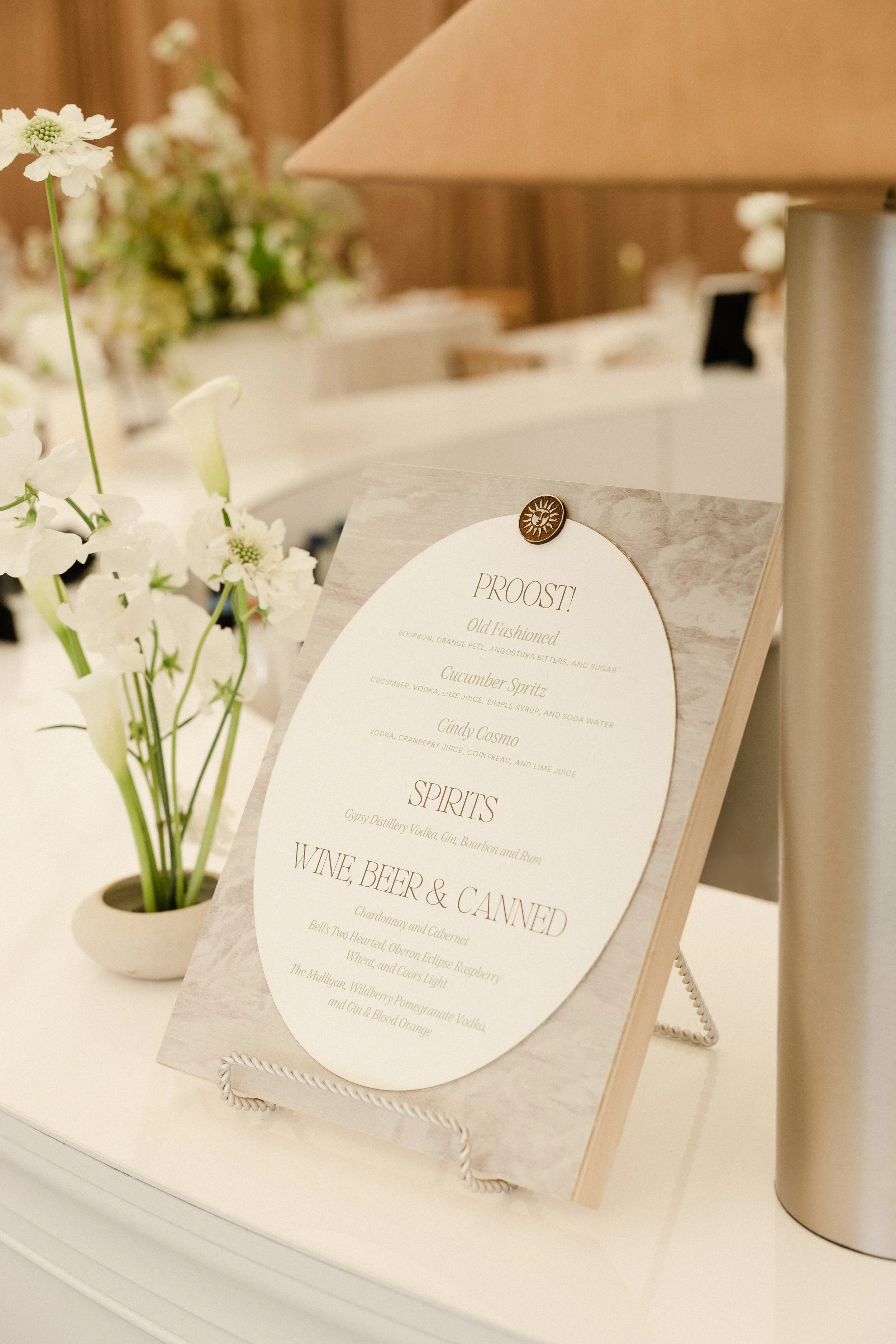 A menu board with drink options and a vase of white flowers on a white counter, with a lamp and other flowers in the background.