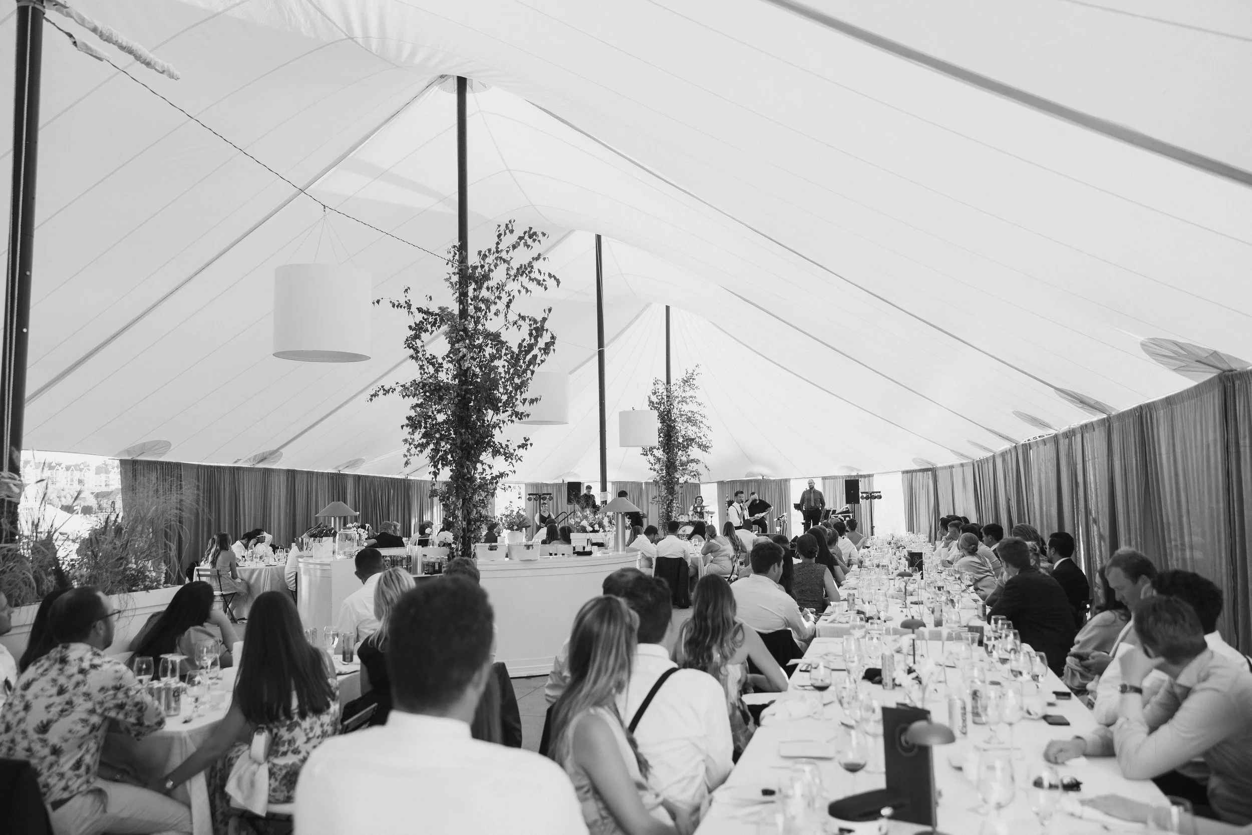 Guests seated at long tables during a wedding reception or banquet under a large tent, with a band or musicians performing in the background.