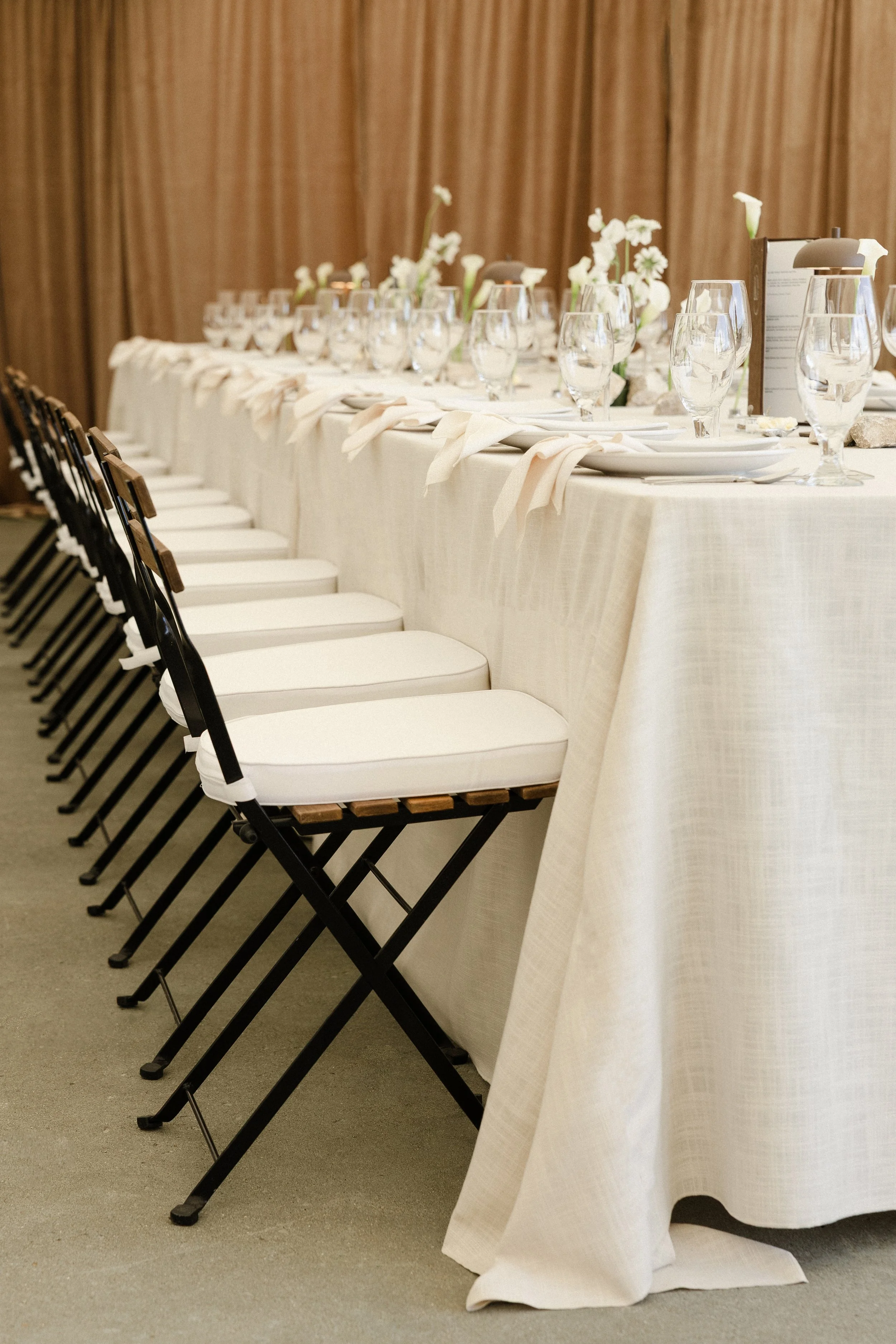Wedding reception table set with white tablecloth, glassware, and floral centerpieces, with black and white chairs arranged along the side.