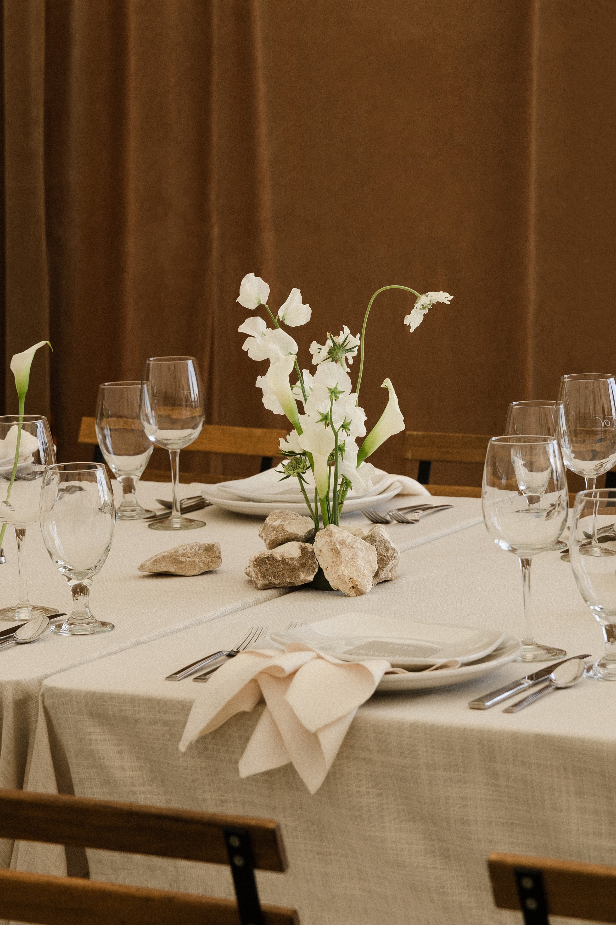 Elegant dining table with a white tablecloth, decorated with a floral centerpiece made of white flowers and stones, surrounded by glassware and silverware, set against a brown curtain background.