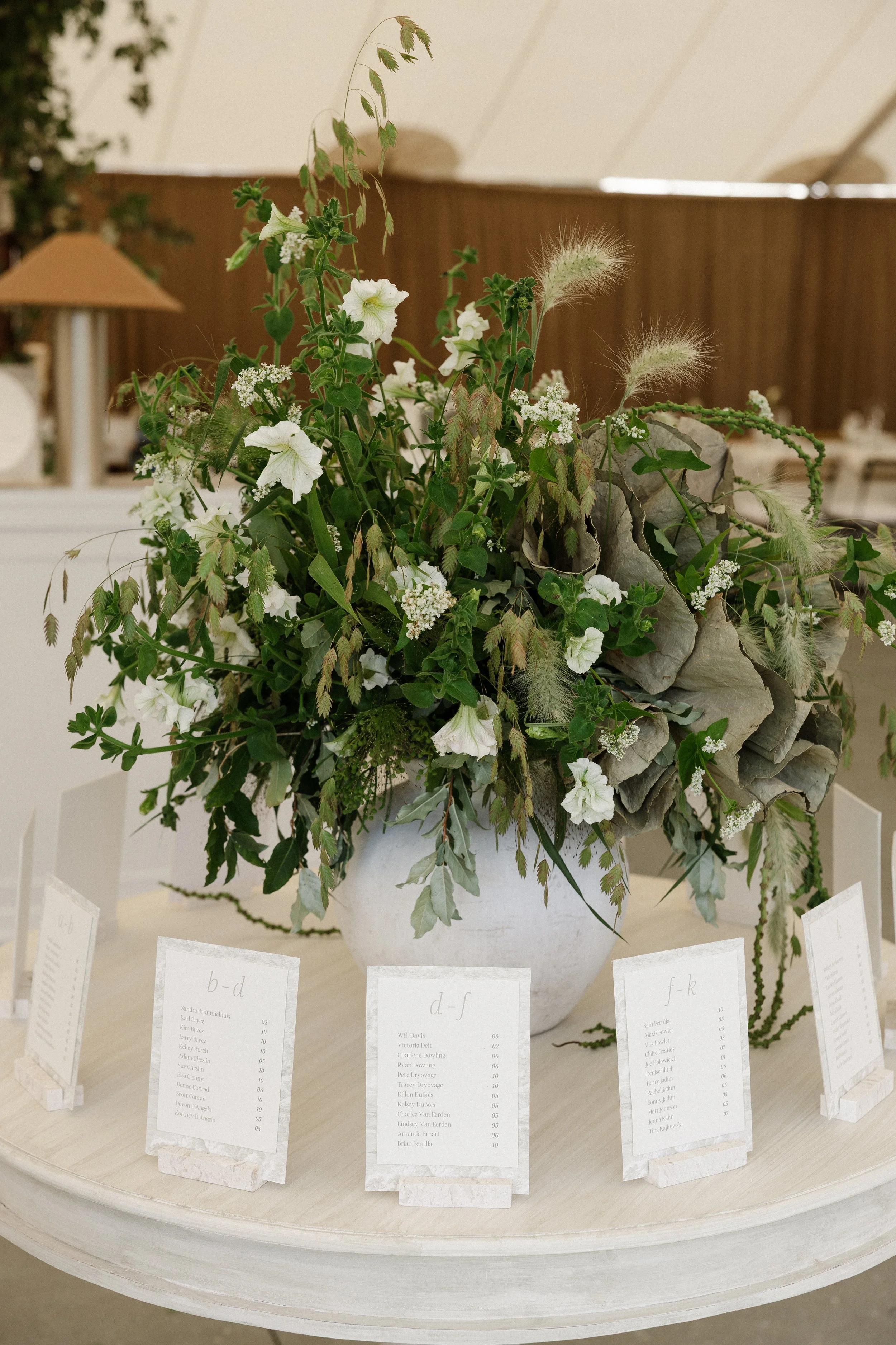 A large white flower arrangement with white flowers, greenery, and soft pampas grass in a white ceramic vase, displayed on a round table with seating cards underneath, in a warm indoor setting.