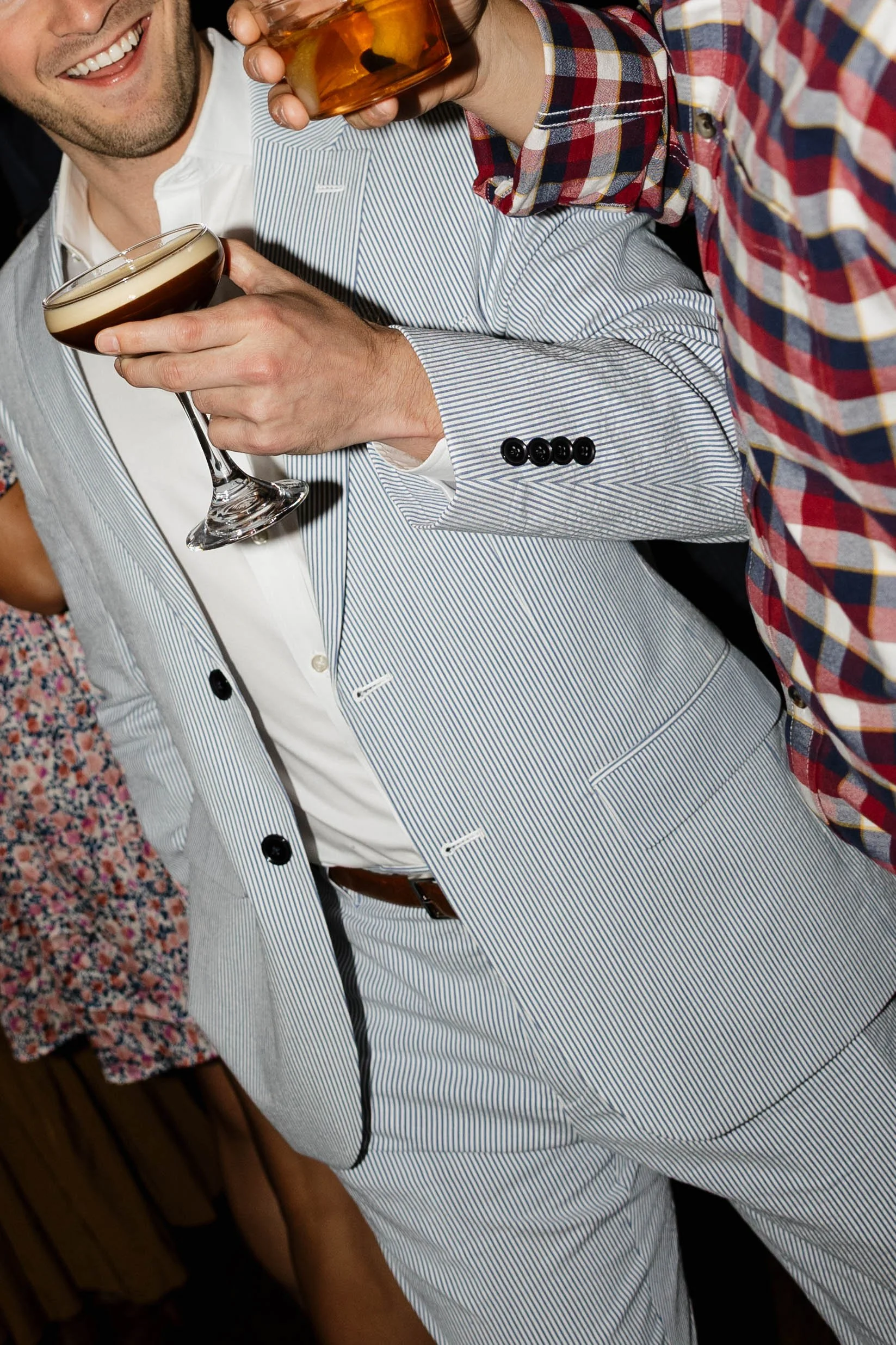 Two men at a party holding drinks, one with a cocktail glass and the other with a whiskey glass, dressed in formal and casual attire respectively.