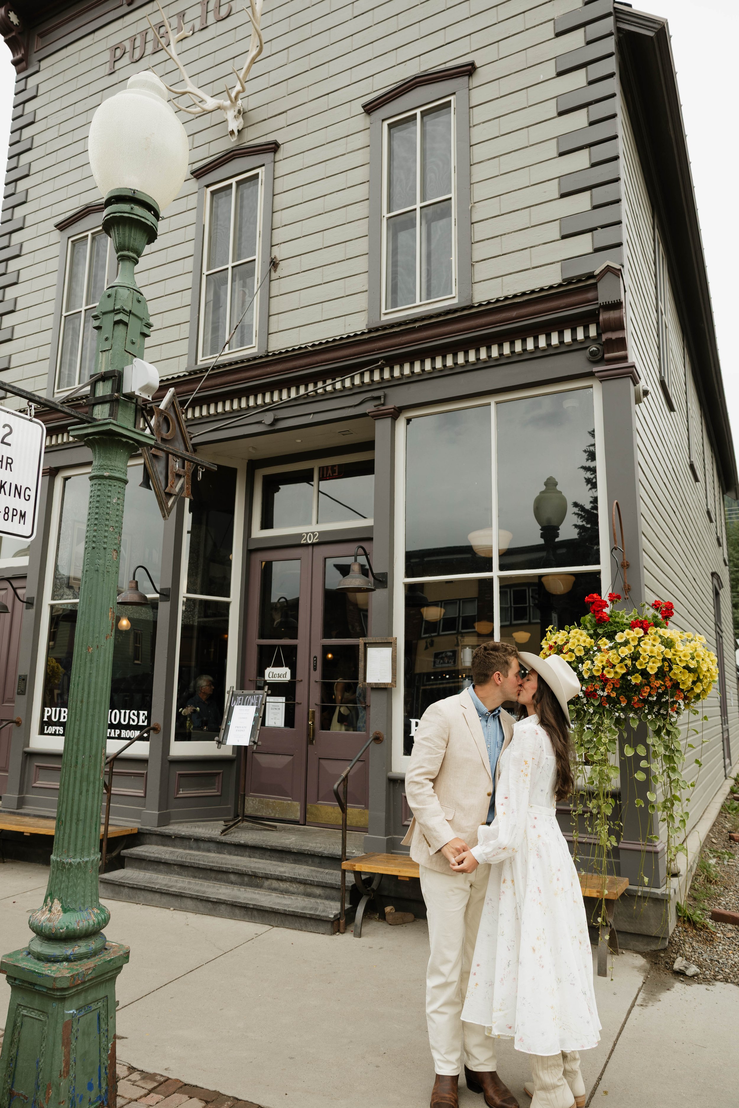 A young couple dressed in vintage-style wedding attire sharing a kiss outside a historic pub with large windows and a flower arrangement in a planter.