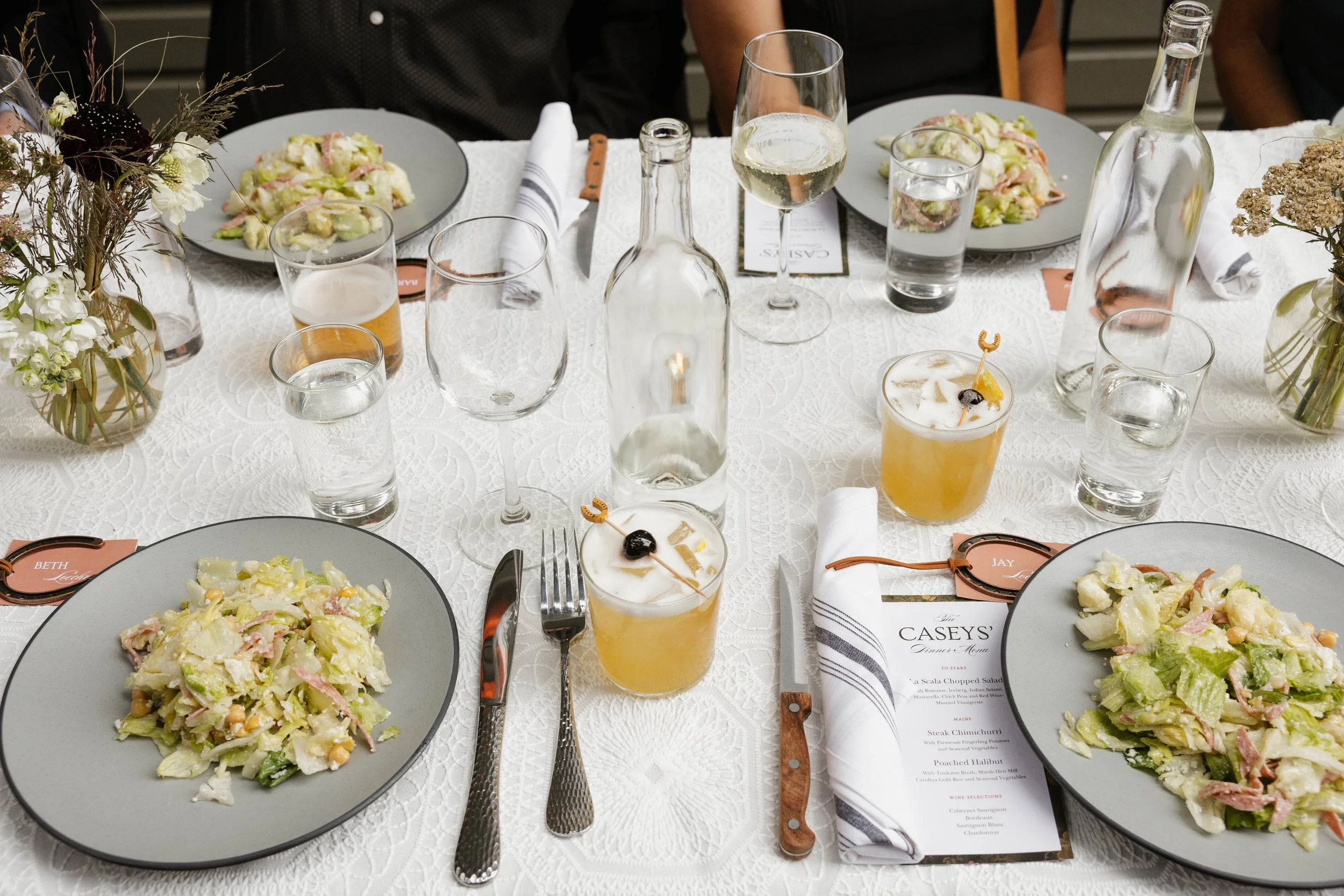A formal dining table set with plates of salad, glasses of water, wine, and cocktails, accompanied by a printed menu, cloth napkins, forks, and vases with flowers.