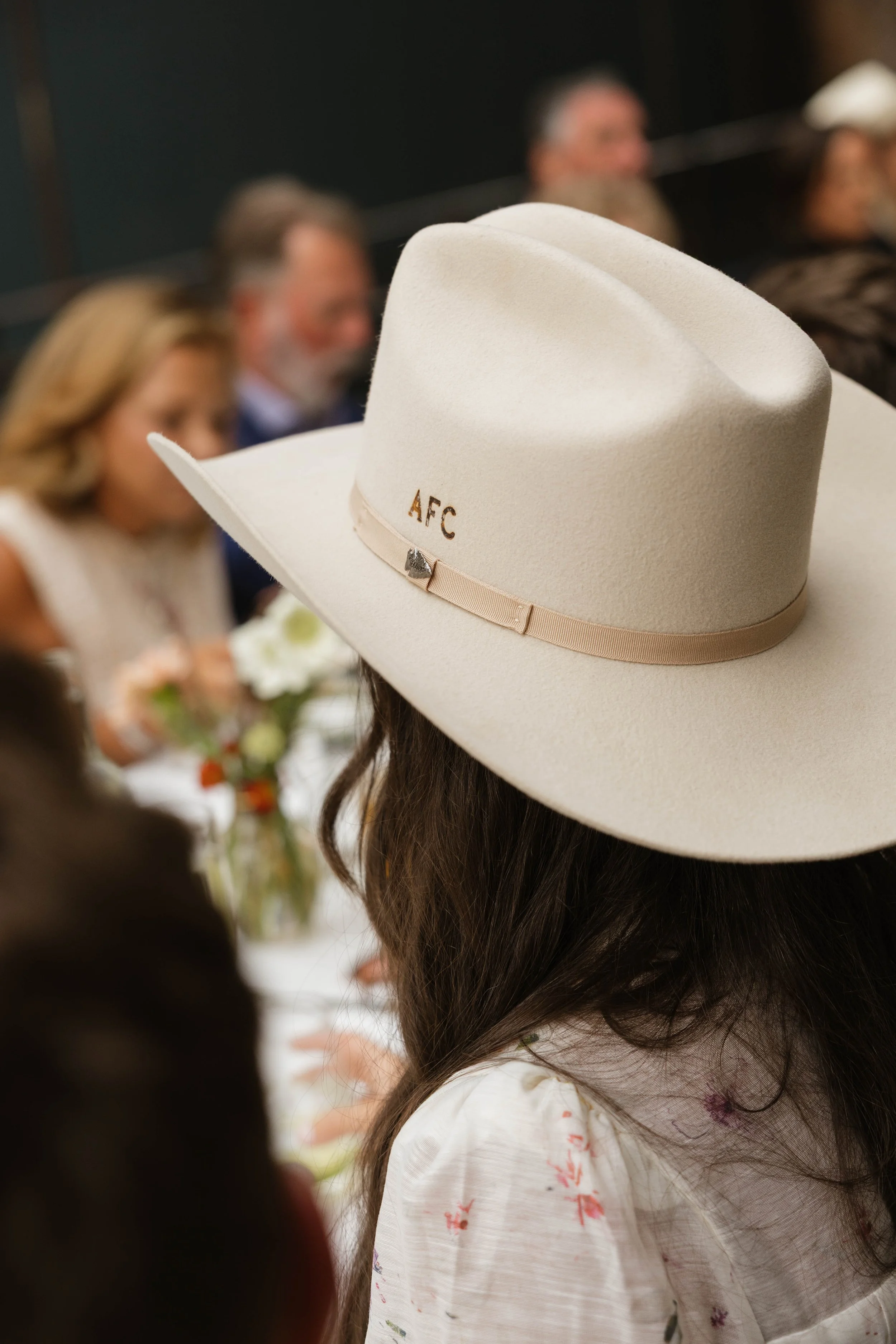 A woman is wearing a large white cowboy hat with the initials 'AFC' embroidered on it, at a social event with people seated at a table in the background.