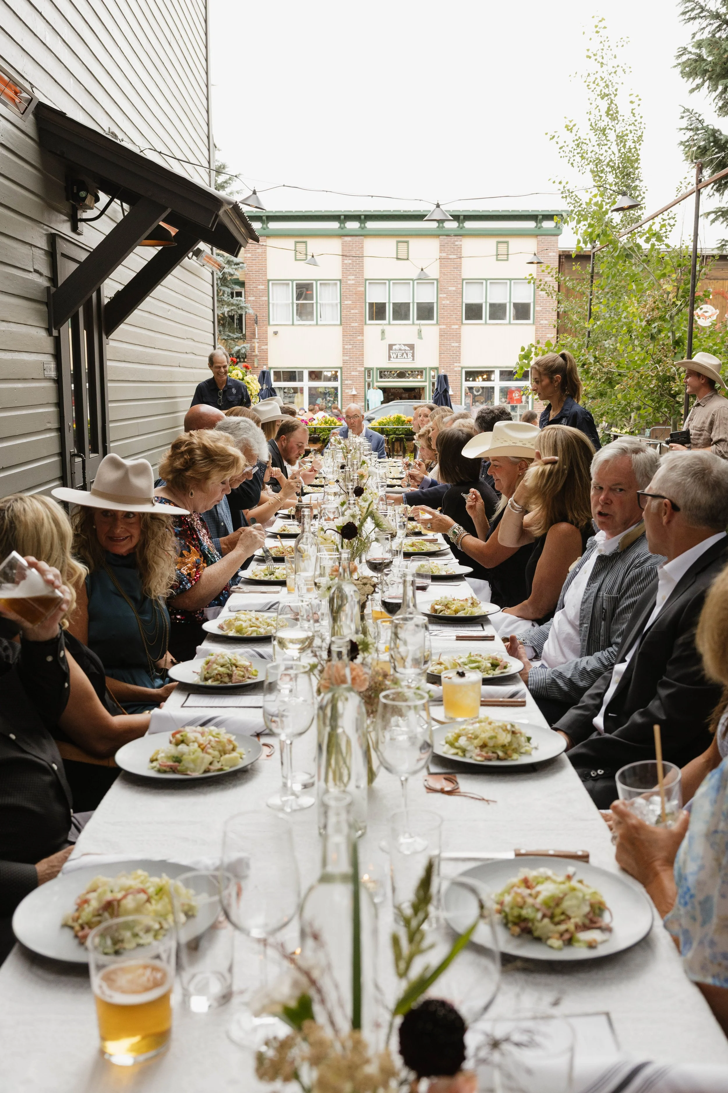 People gathered at a long outdoor dining table enjoying a meal during a social event in a city setting with buildings and trees in the background.