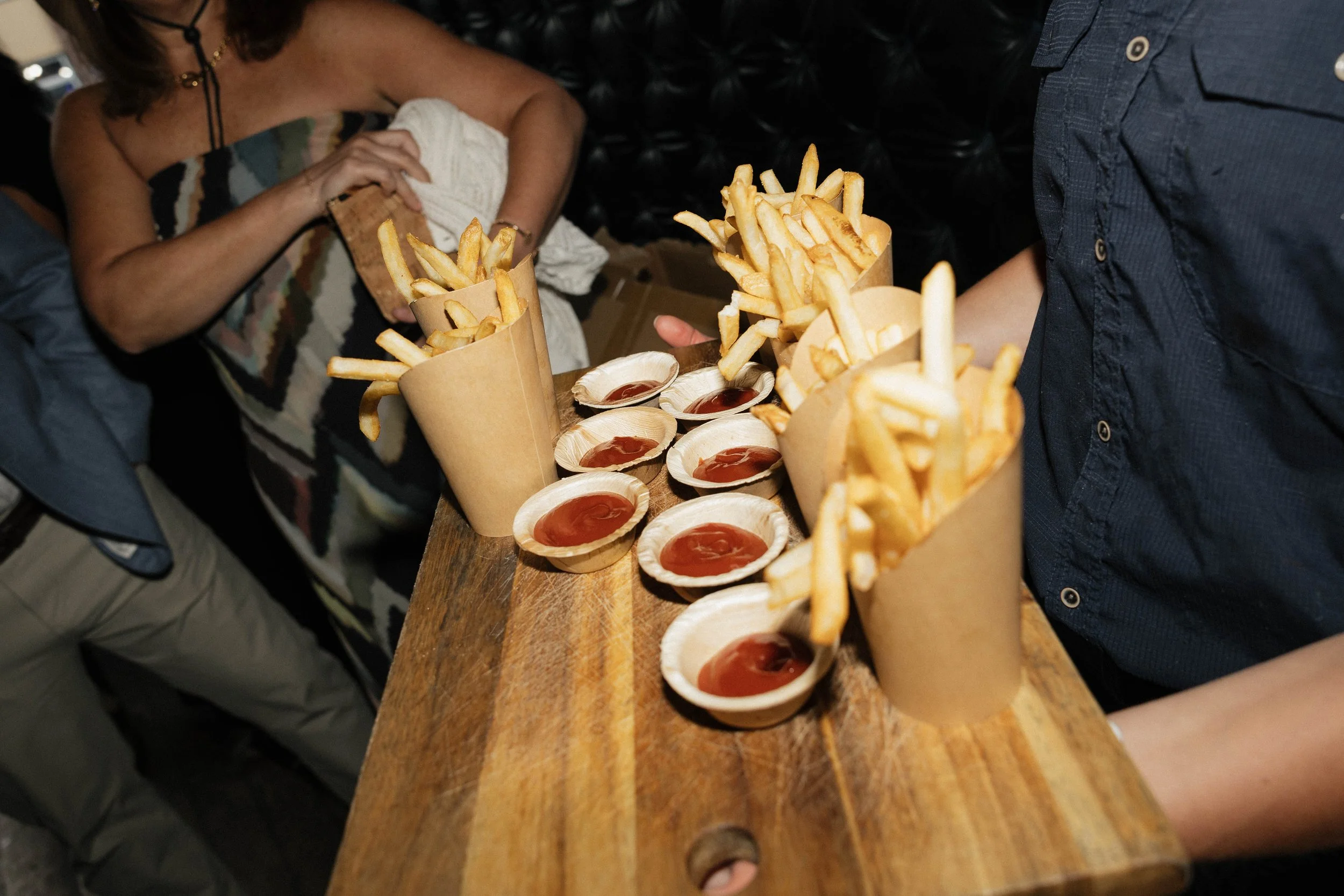 French fries in paper cones with small dishes of ketchup on a wooden table in a casual dining setting.