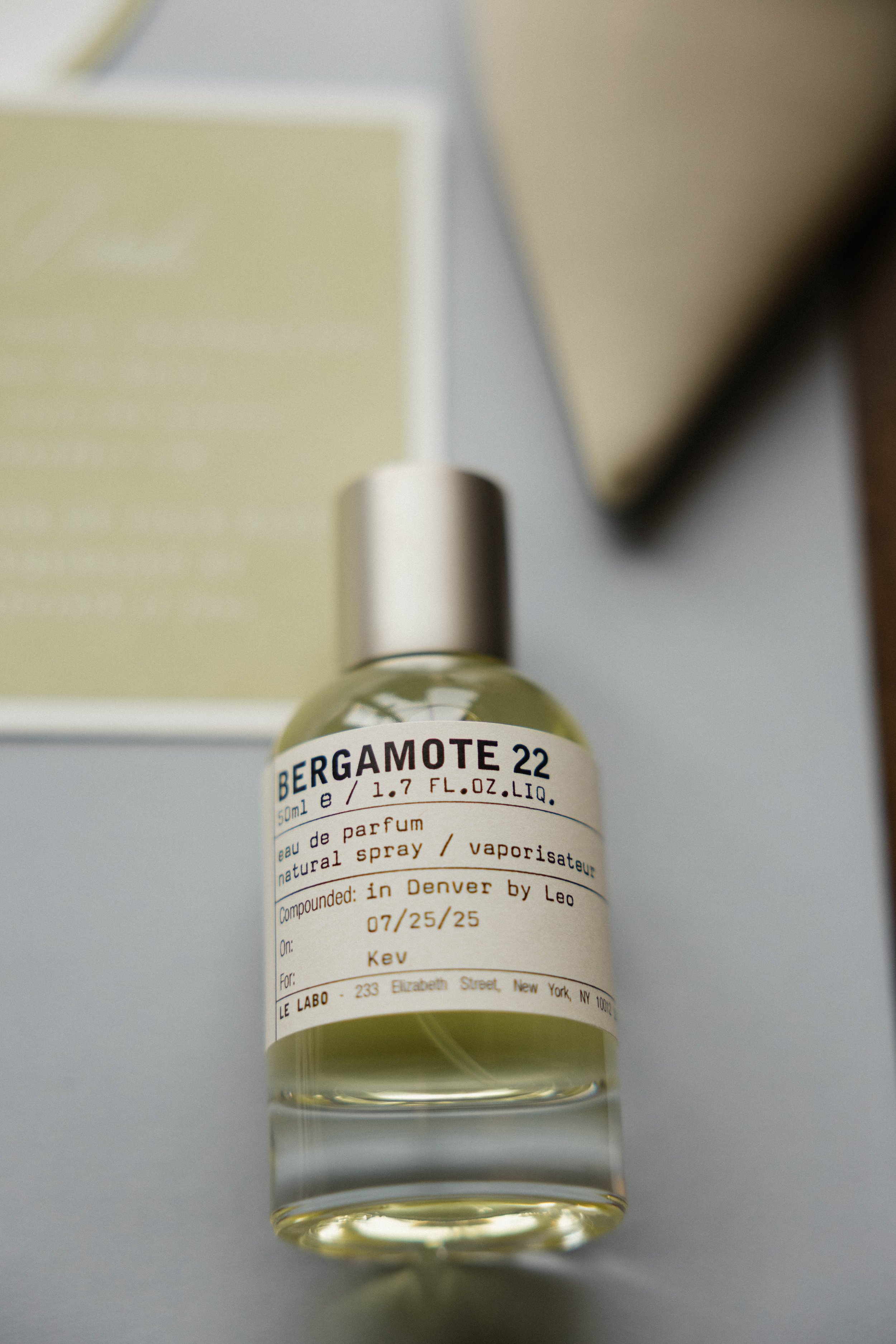 A small glass bottle of Bergamote 22 perfume with a white label and black text, sitting on a light-colored surface with a blurred background.