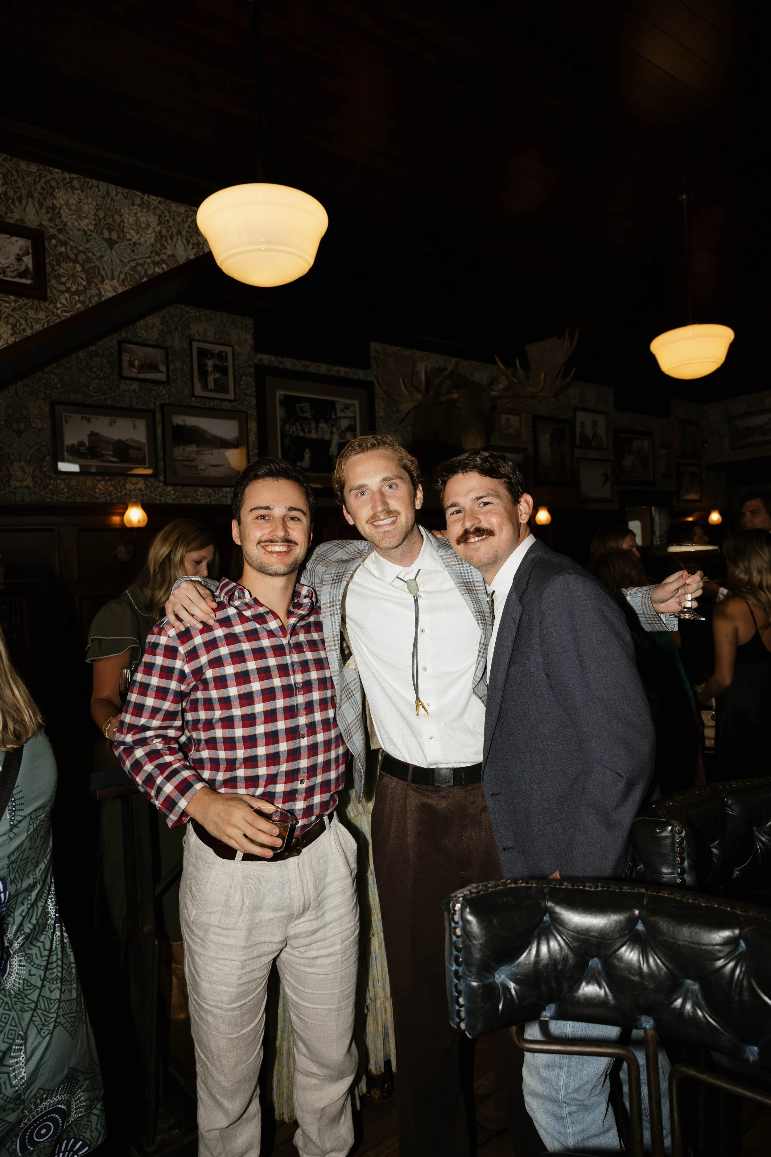 Three men posing together at a social gathering or party, smiling, with friends in a dimly lit bar or restaurant.