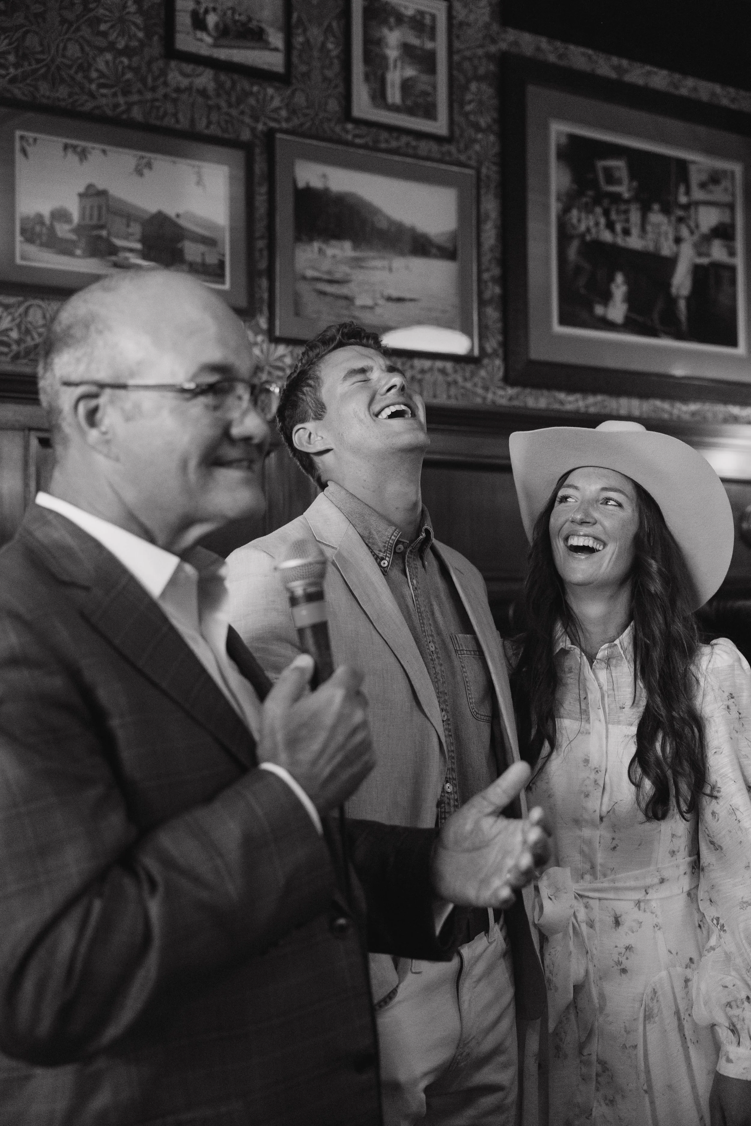 Three people at a social event, with one person holding a microphone, all laughing and enjoying themselves in a room with framed pictures on the wall.