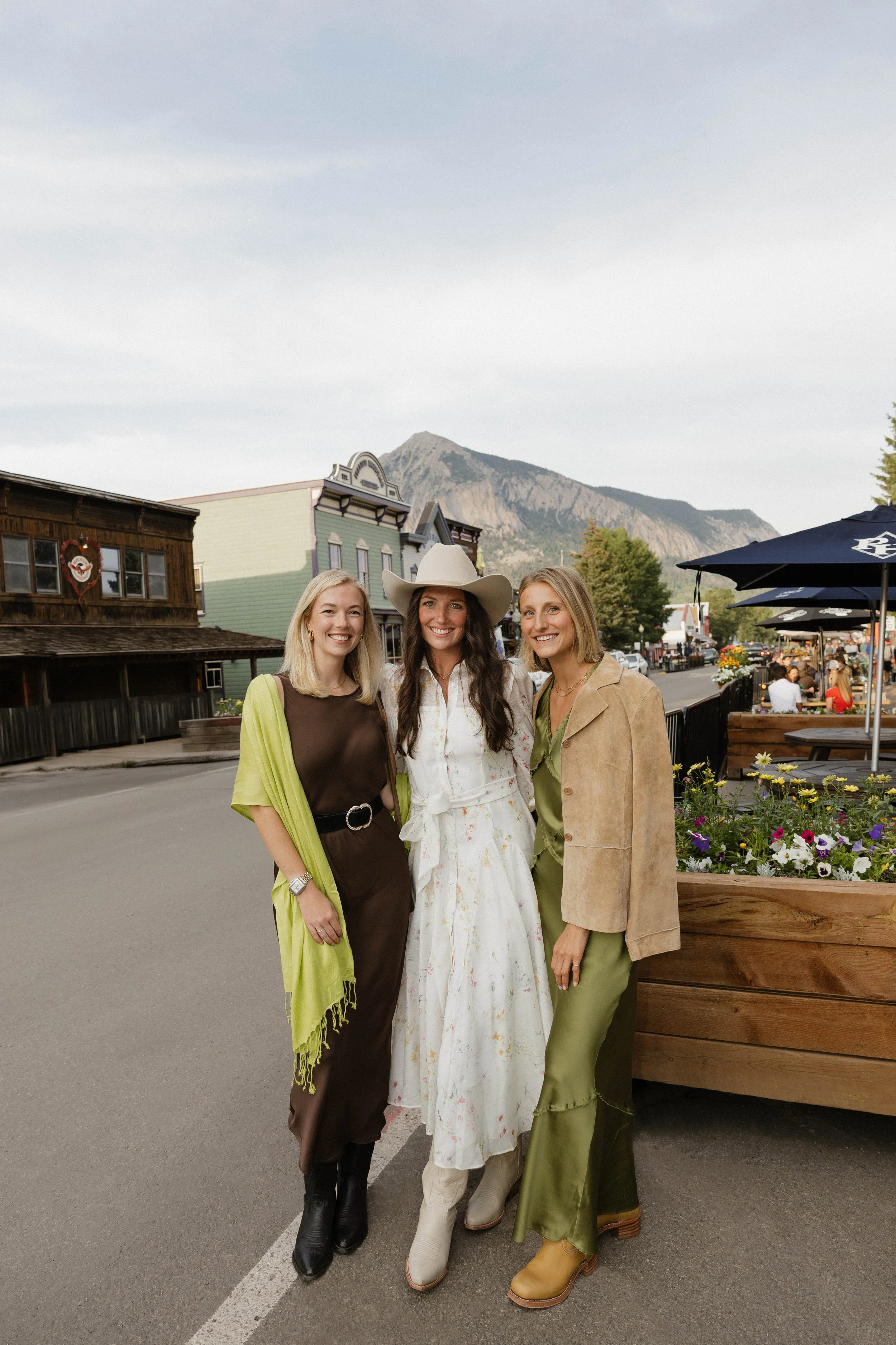 Three women standing together outdoors in a small town, smiling, with mountains in the background.
