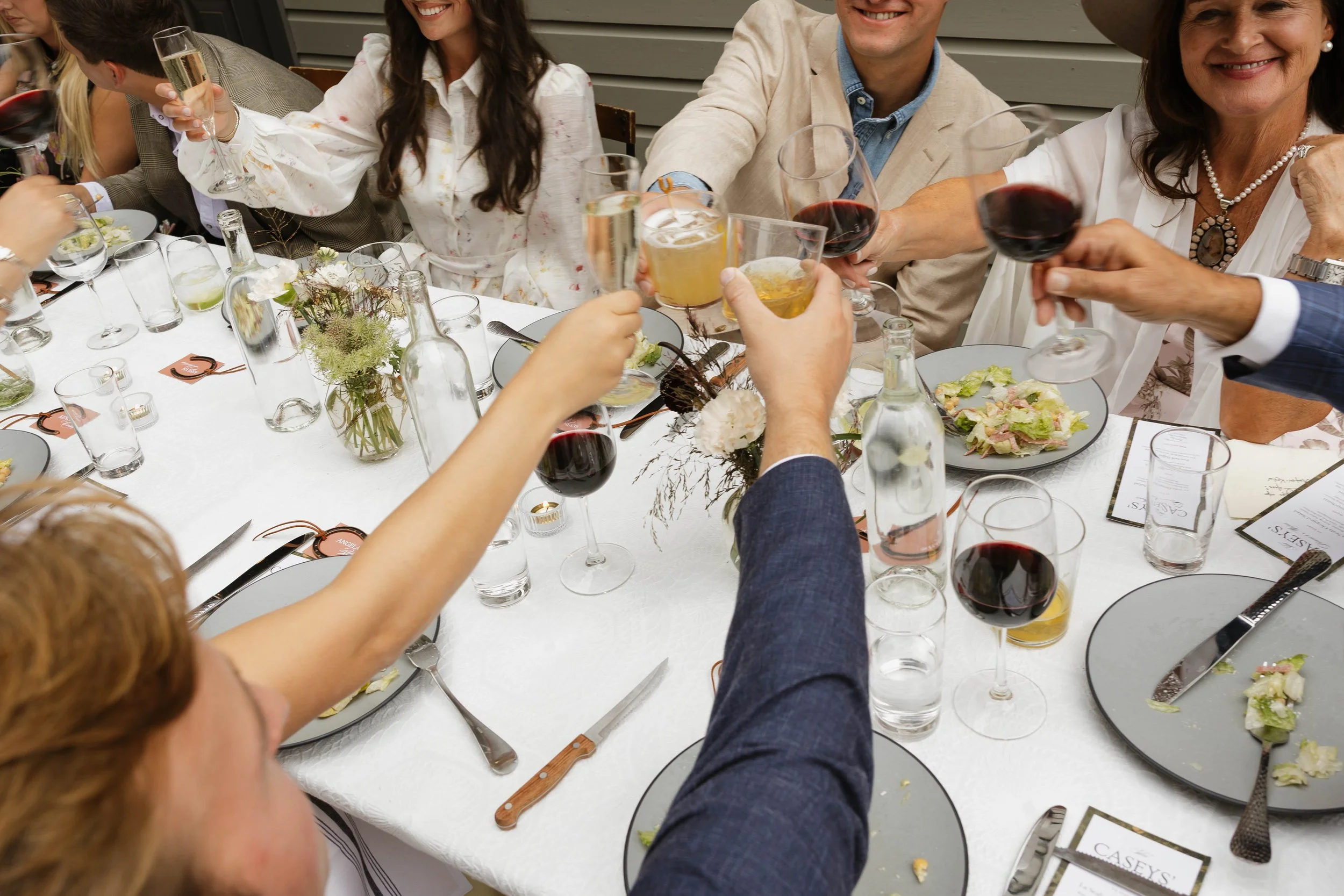 People raising glasses in a toast at a dinner party with a white tablecloth, floral centerpiece, plates, and utensils.