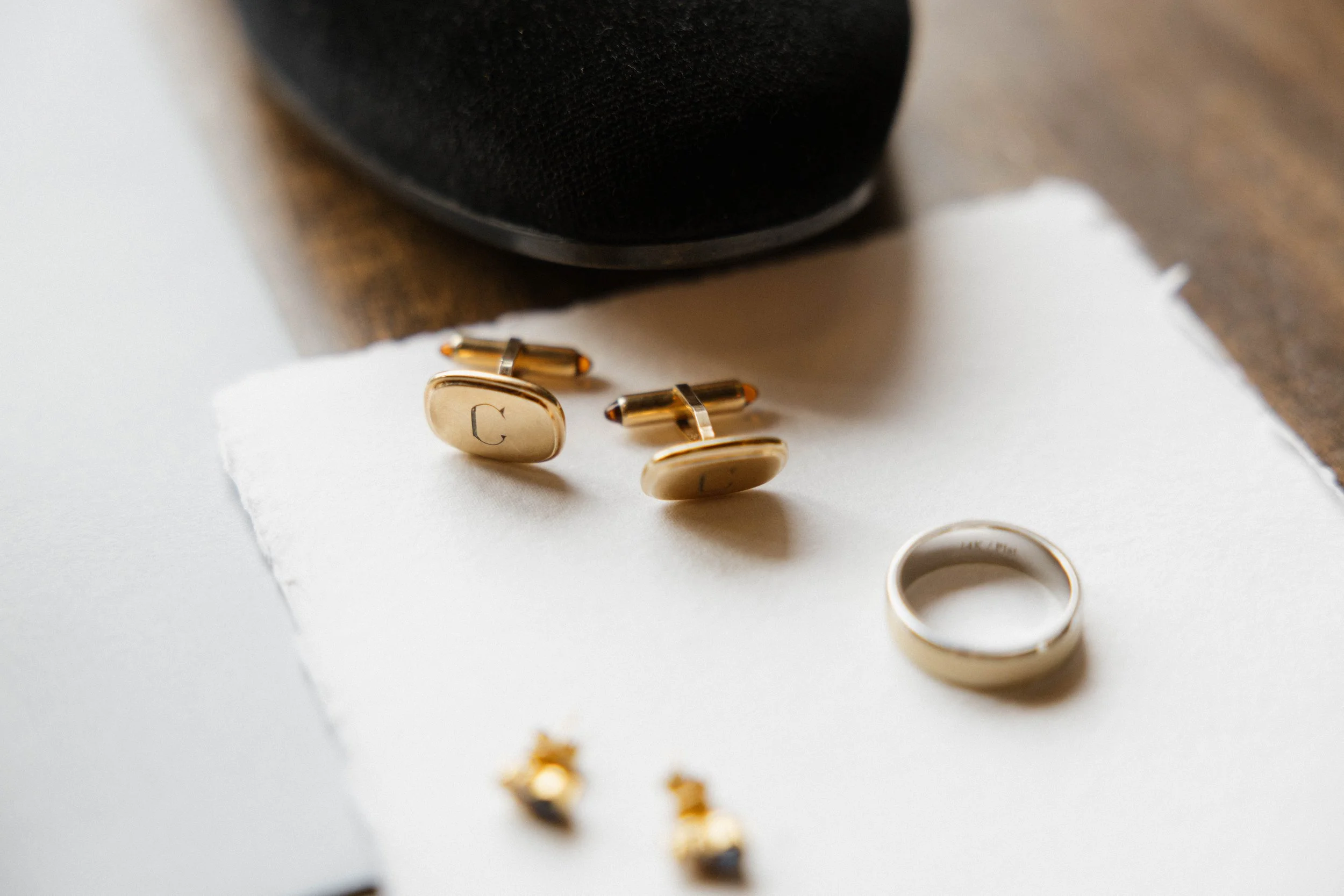Gold cufflinks and a ring on a white piece of paper, with a black box and a wooden surface in the background.
