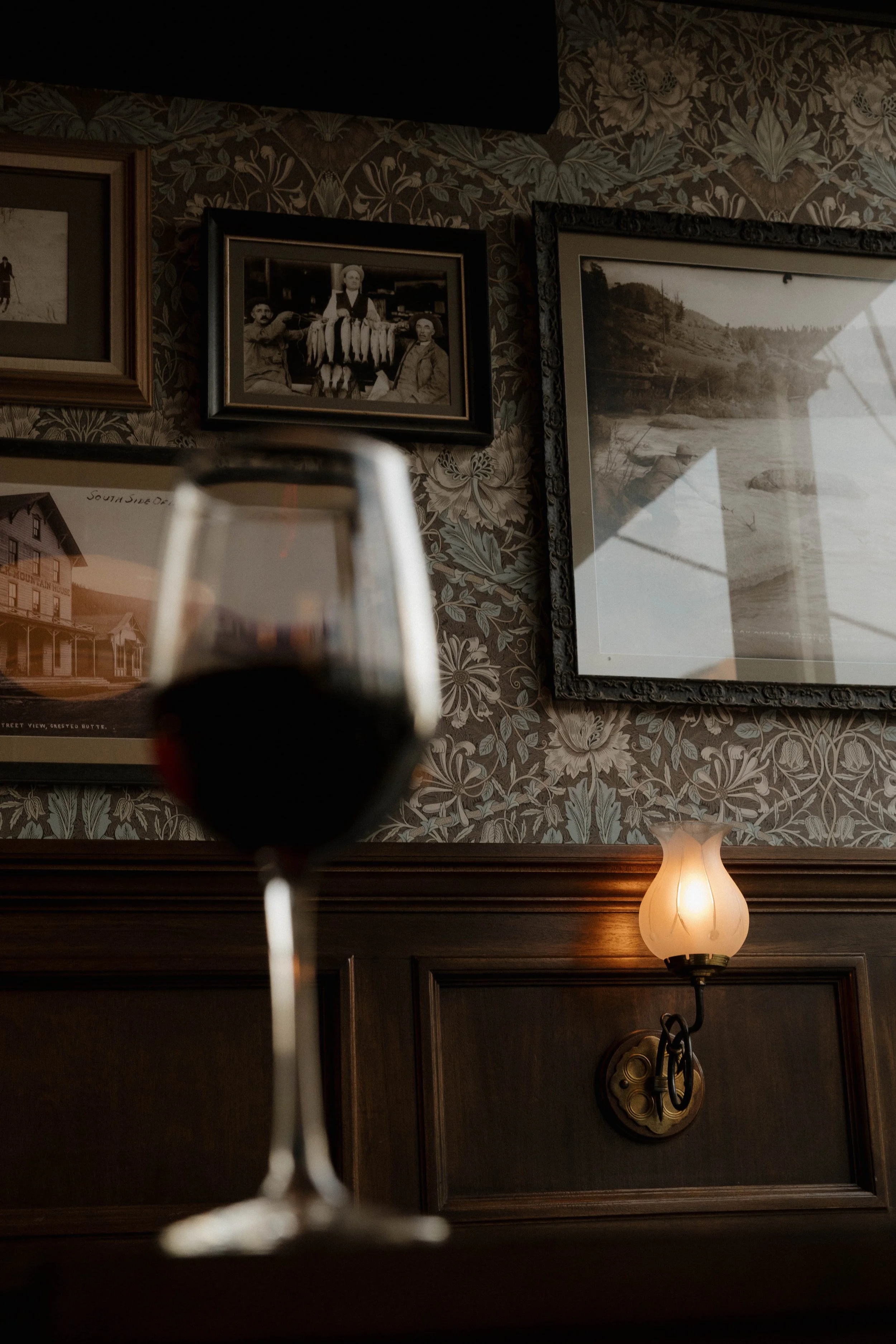 A glass of red wine in the foreground with a dimly lit lamp and framed photographs on a wall in the background.