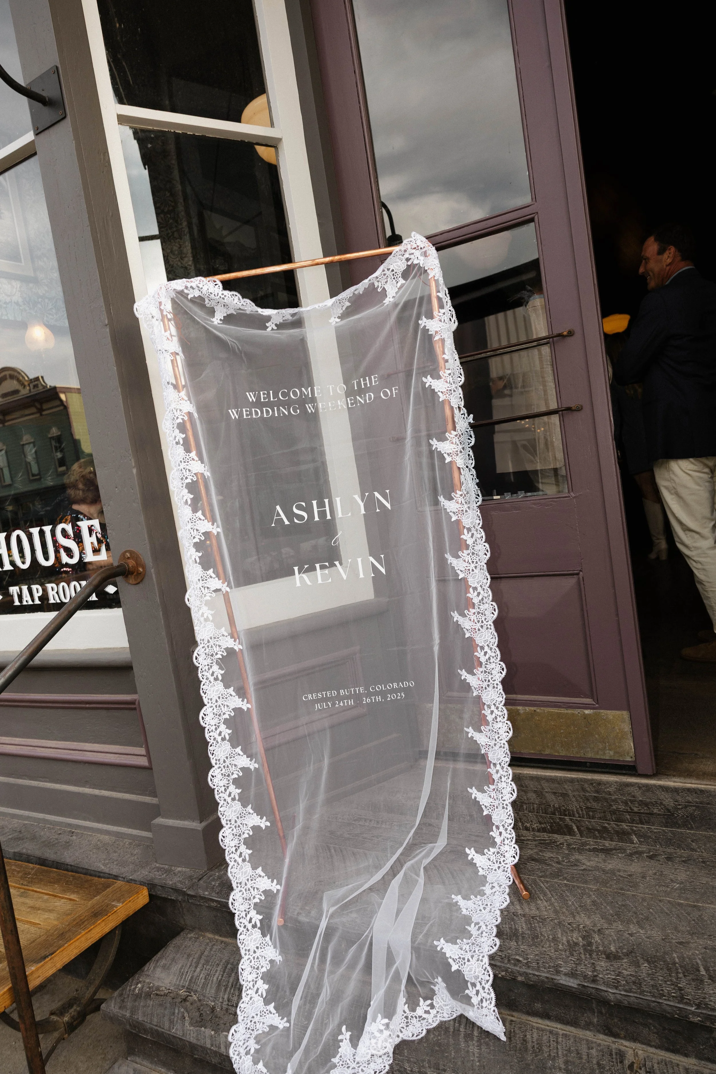 Wedding welcome sign on sheer fabric with lace border outside of a building, indicating the wedding weekend of Ashlyn and Kevin in Crested Butte, Colorado, July 24th-26th, 2023.