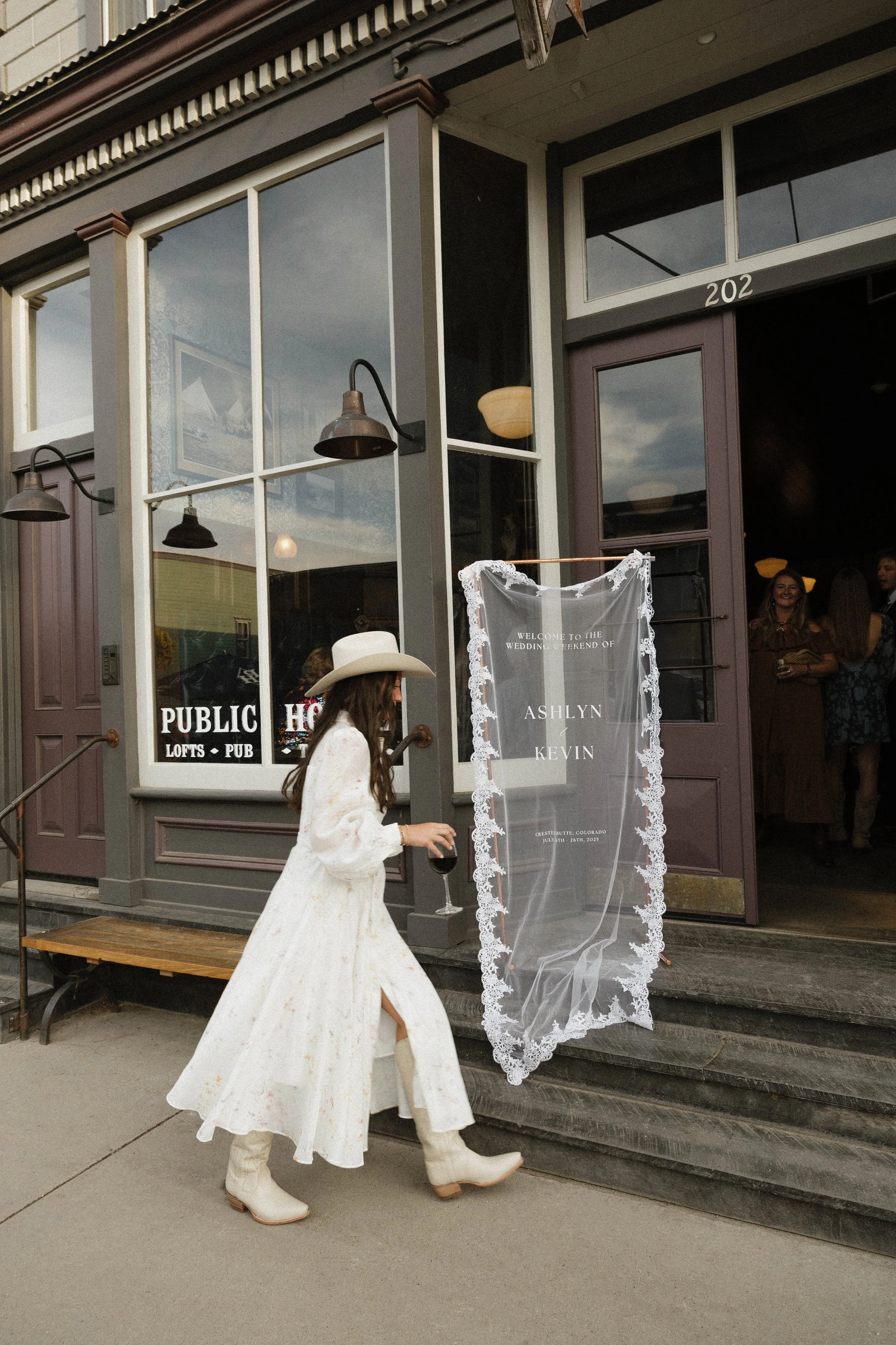 Woman in a white floral dress and cowboy hat walking out of a building holding a wine glass. The building's exterior features large windows and signs that read 'PUBLIC HOUSE LOFTS PUB' and a sign on the door with the names Ashlyn and Kevin, indicating a wedding event.