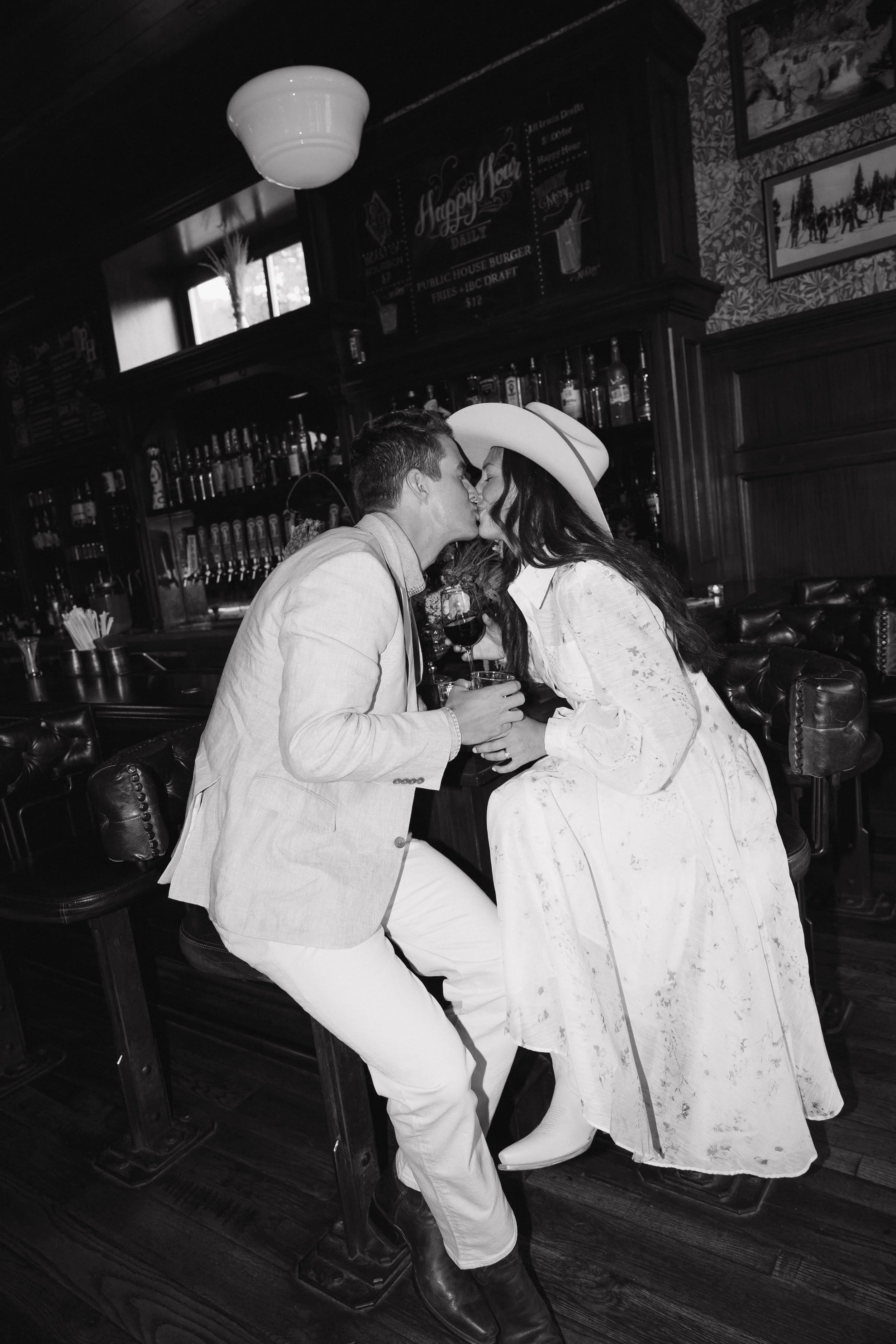 A black-and-white photo of a couple kissing inside a bar, with the woman in a long dress and hat, and the man in a suit jacket, both holding drinks.