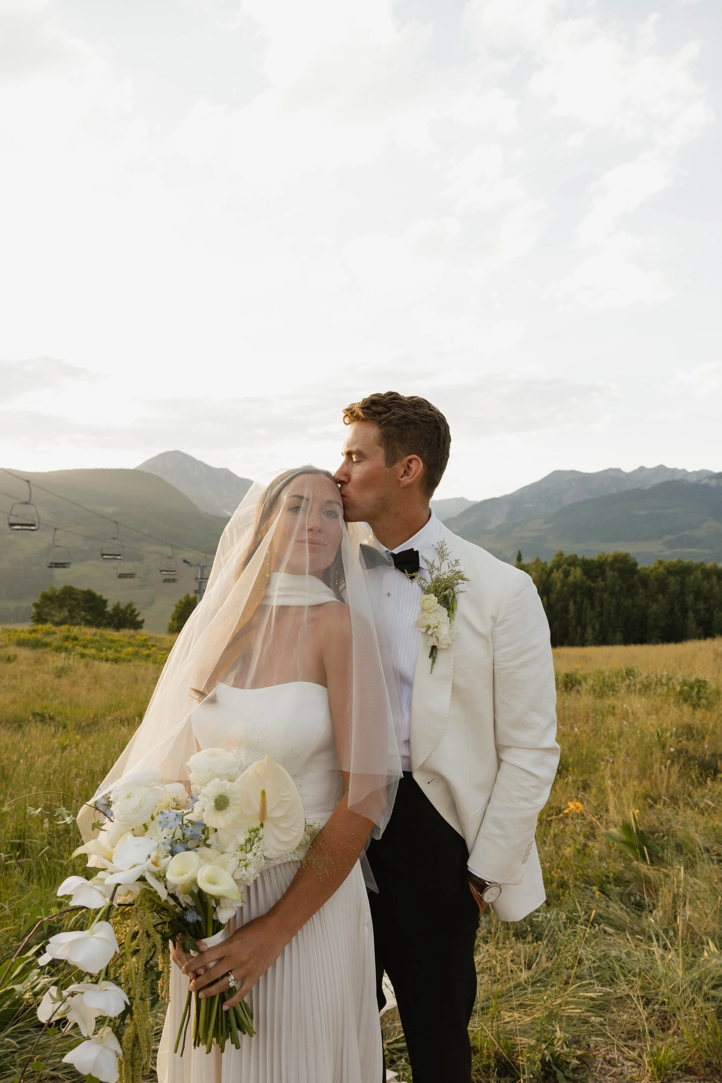 A bride and groom share a kiss in a mountain meadow during their wedding day, with a backdrop of mountains, greenery, and a cloudy sky.