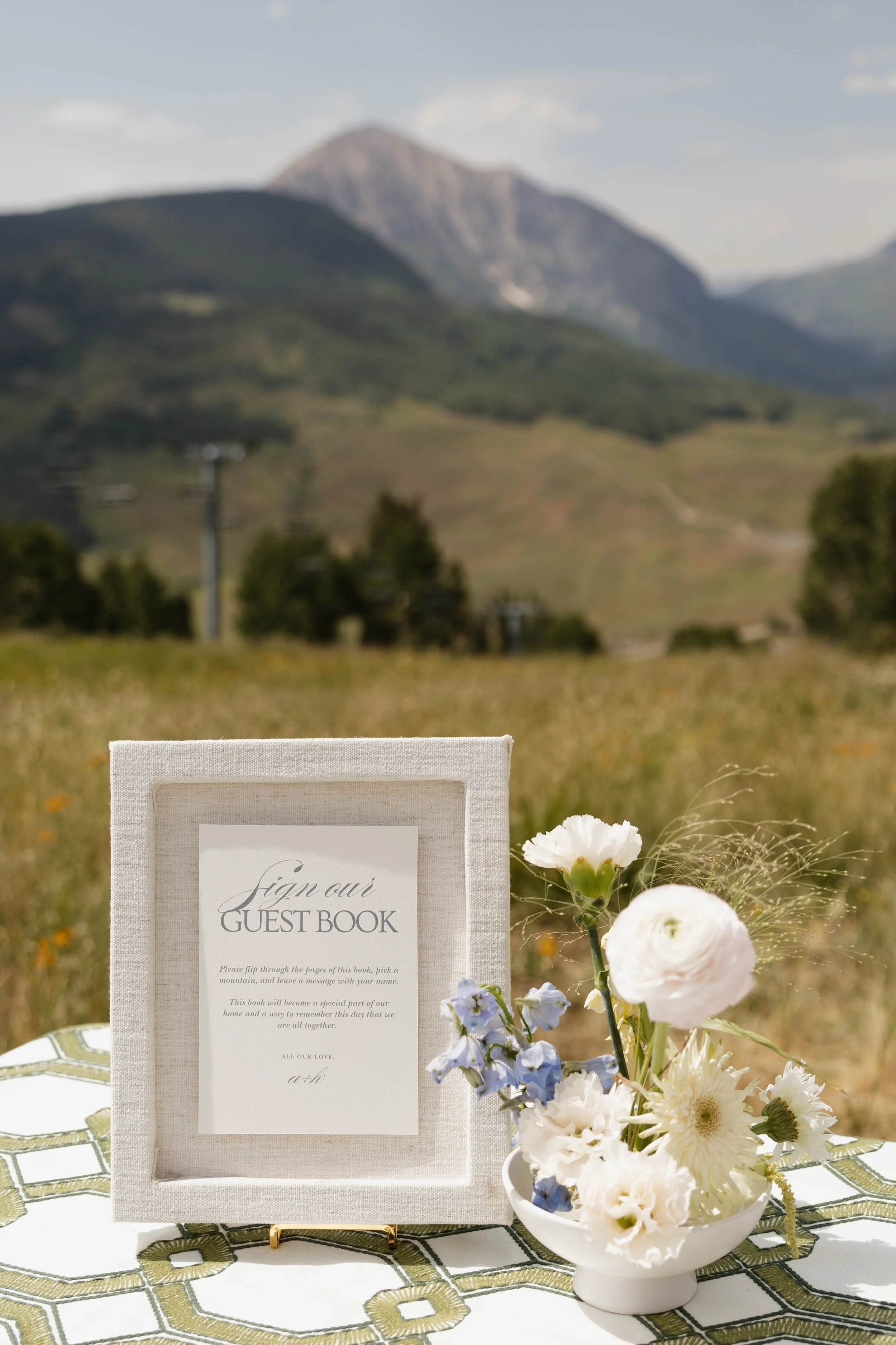 A guest book table with a framed sign and a flower arrangement outdoors with mountains and a field in the background.