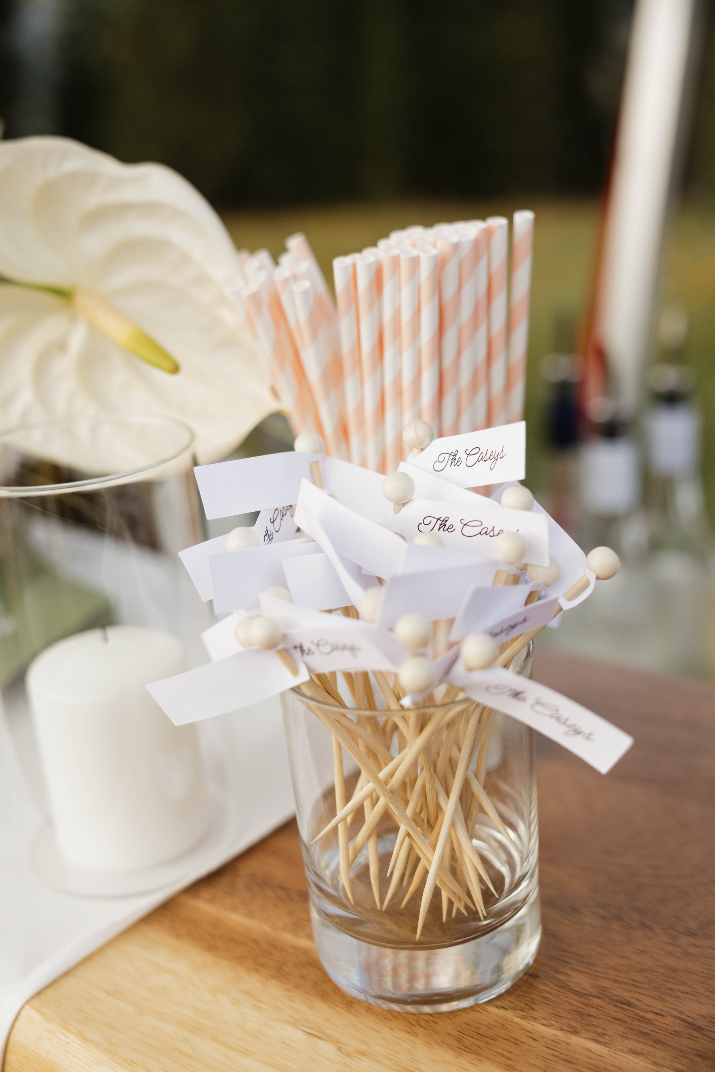 A glass jar filled with white lollipop sticks with paper flags labeled 'The Casbys.' Behind it are pink and white striped paper straws and a container with large white flowers and a white candle.