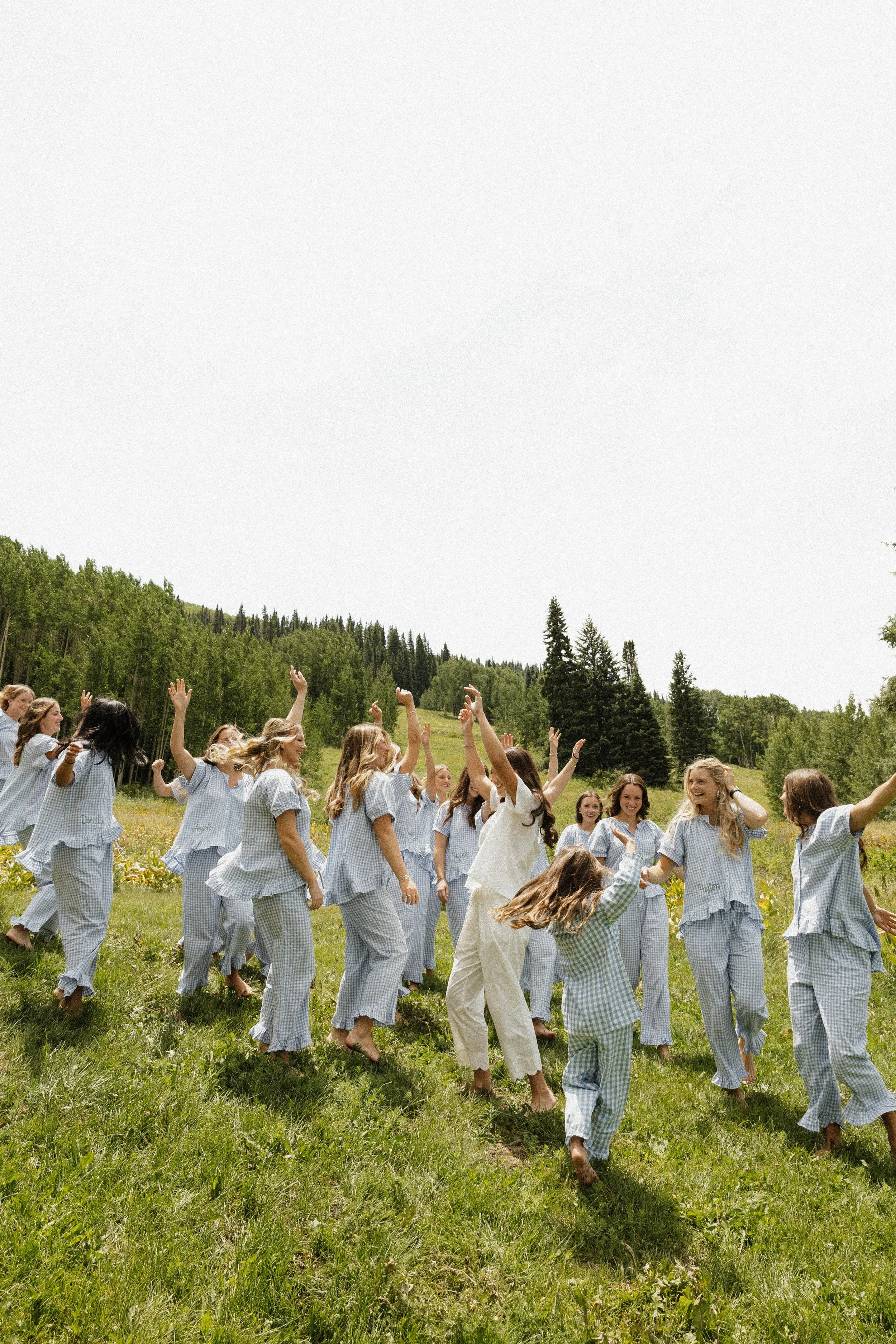 Group of women and girls in matching pajamas laughing and dancing outdoors in a grassy field with trees and hills in the background.