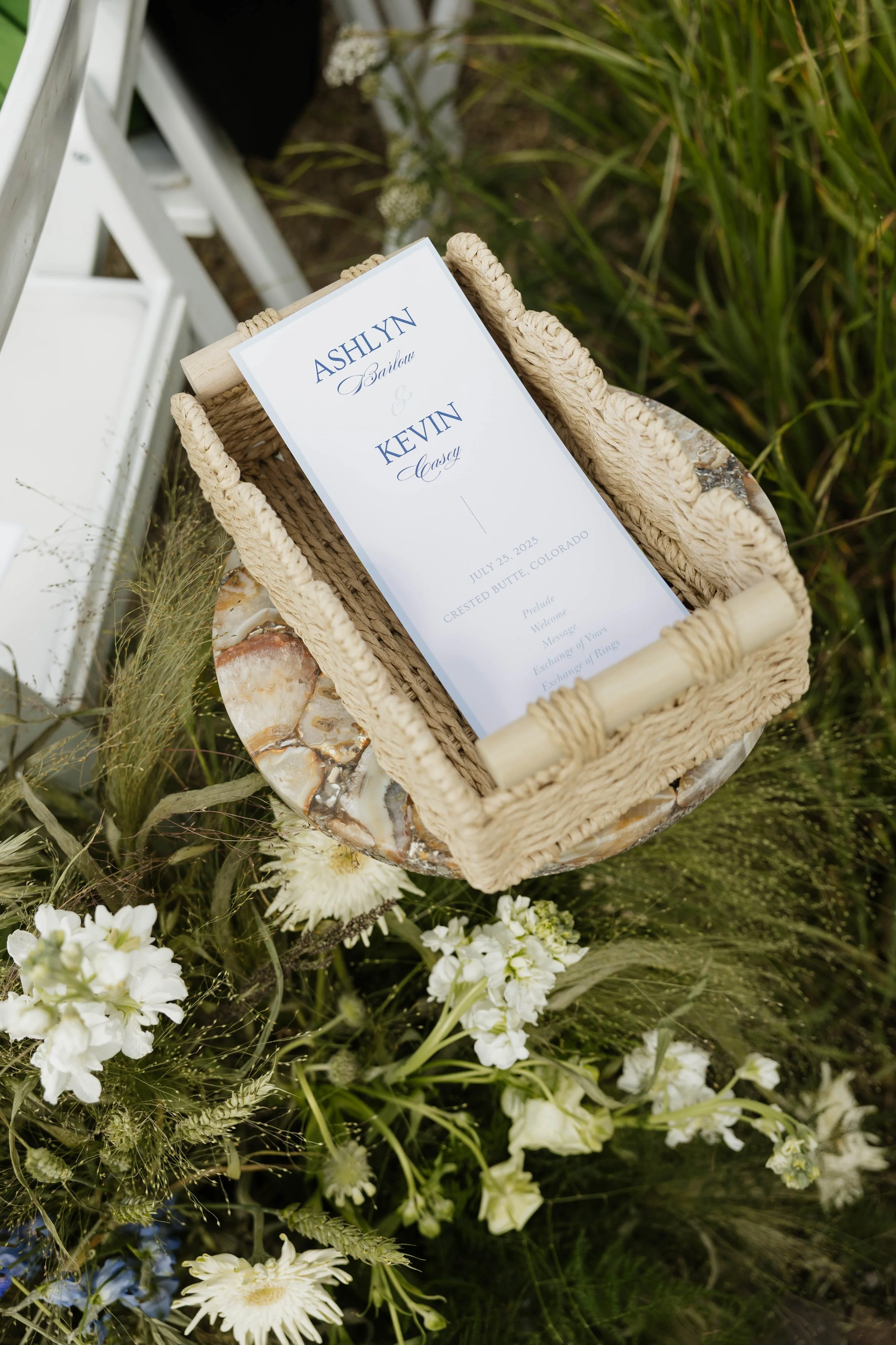 Wedding program card in a woven basket on a small table surrounded by white flowers and greenery.