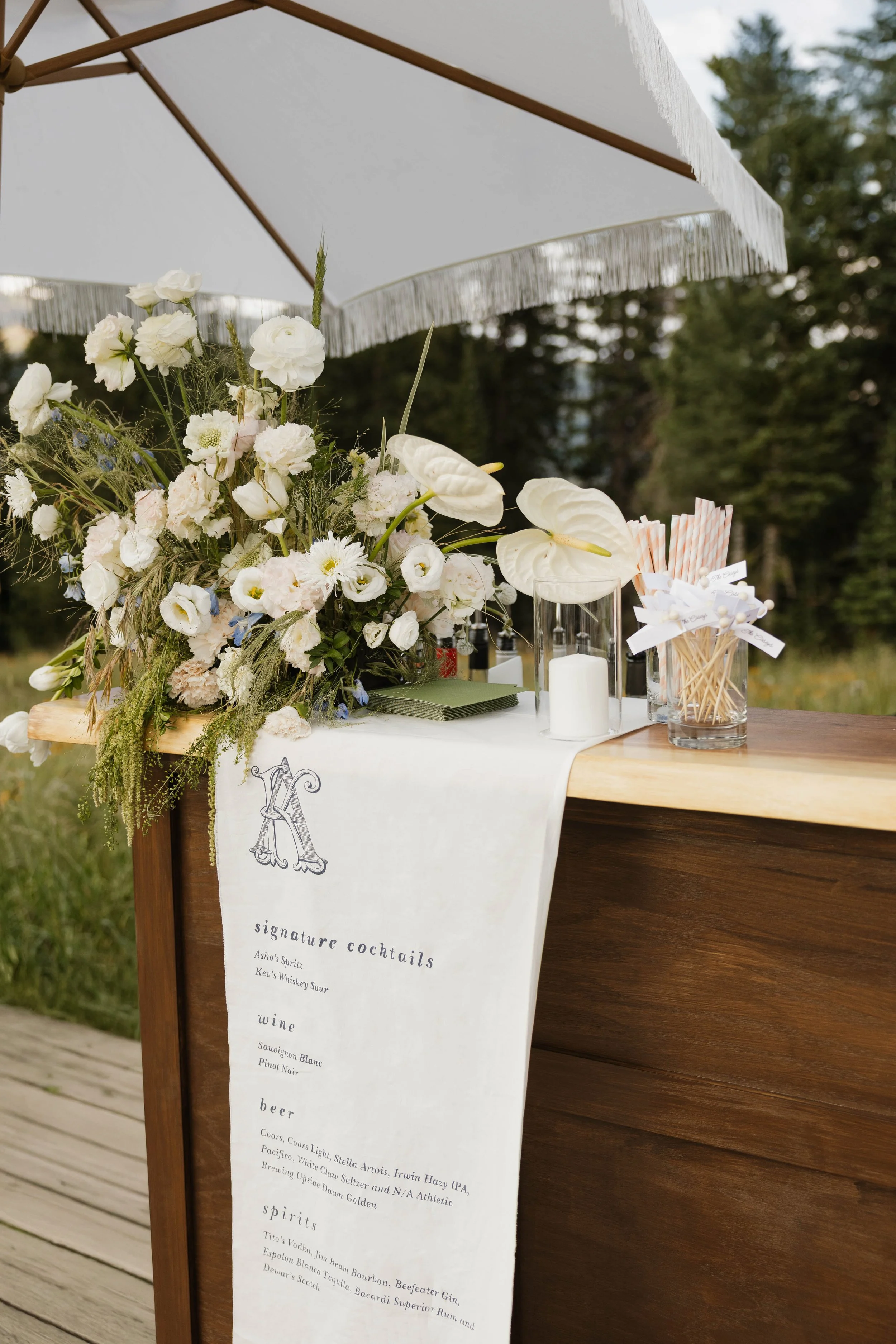 A wedding or event bar setup with a white umbrella, floral arrangements, candles, and a printed menu hanging off the edge of a wooden bar, outdoors with trees in the background.