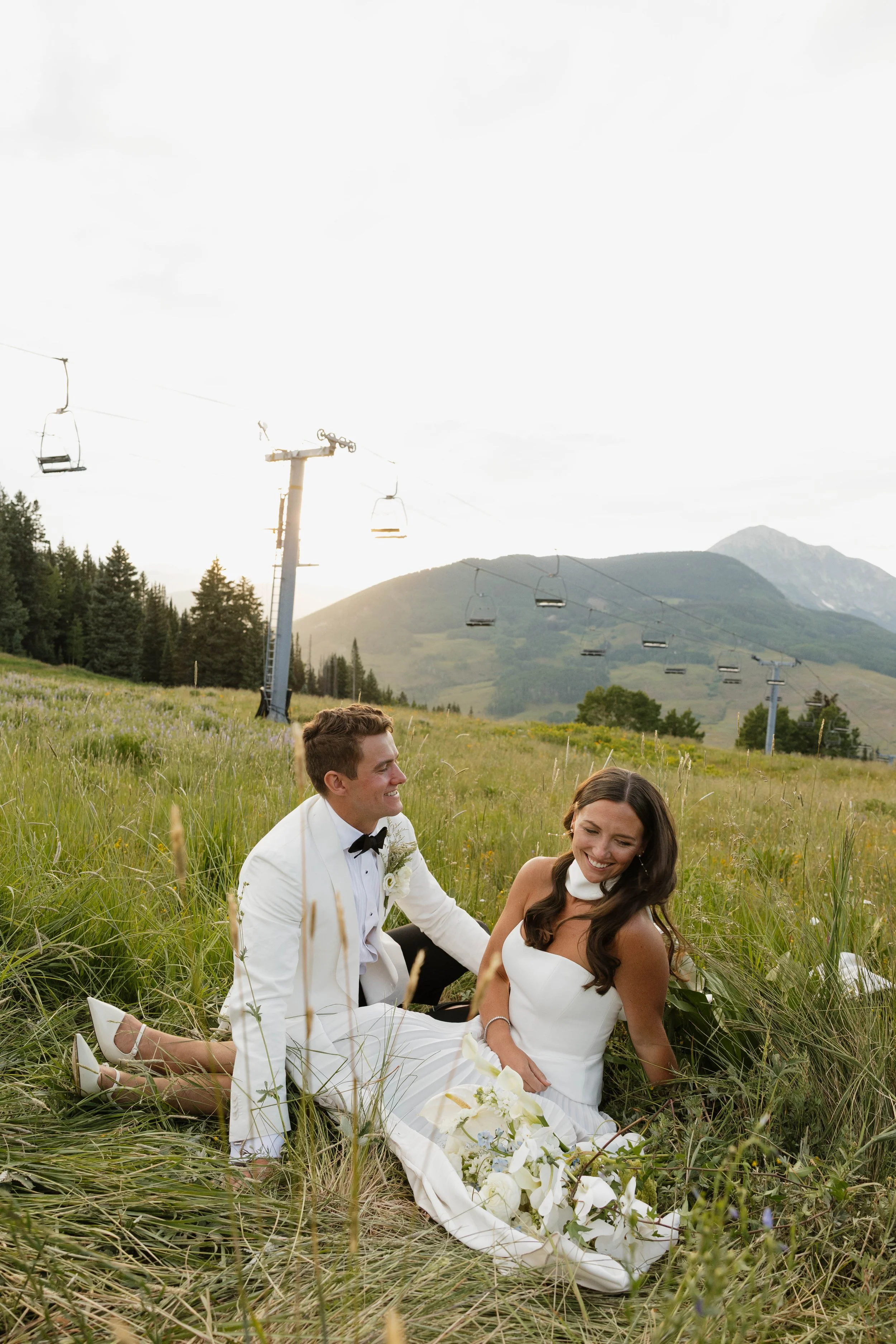 A newlywed couple sitting on grass in a field, smiling and looking at each other, with ski lift chairs and mountains in the background during daytime.