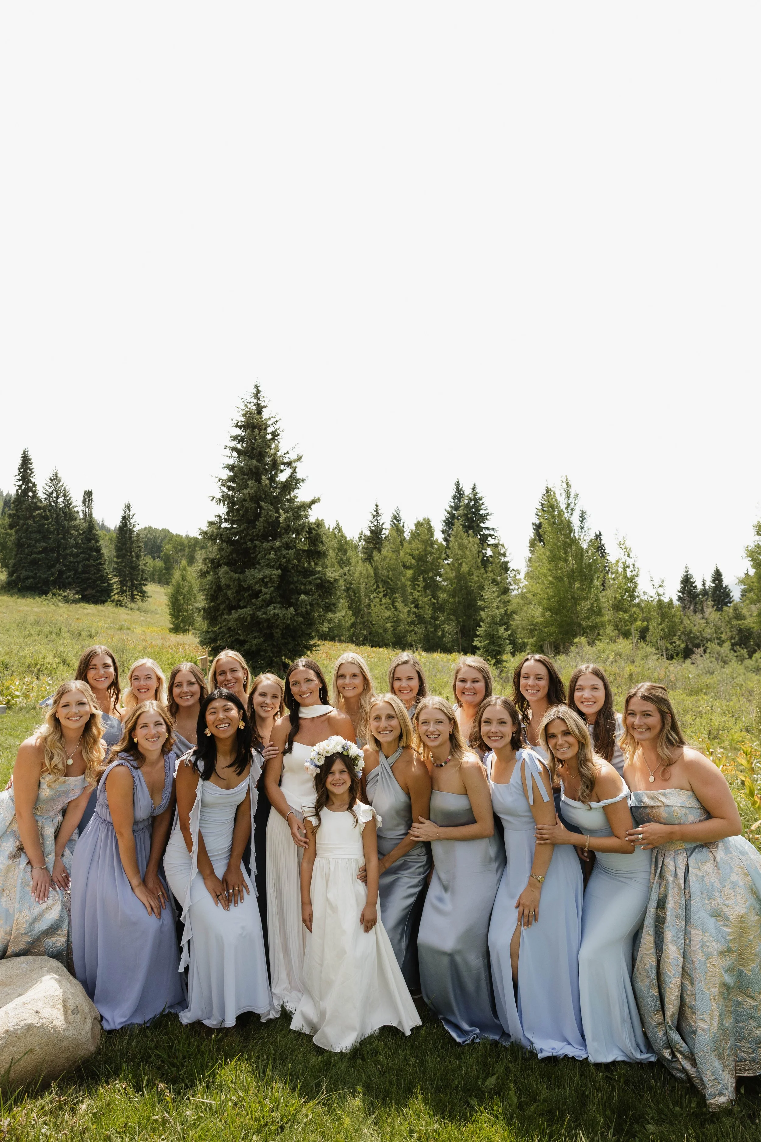 Group of women in light blue dresses and a young girl in a white dress posing outdoors in a green field with trees in the background.