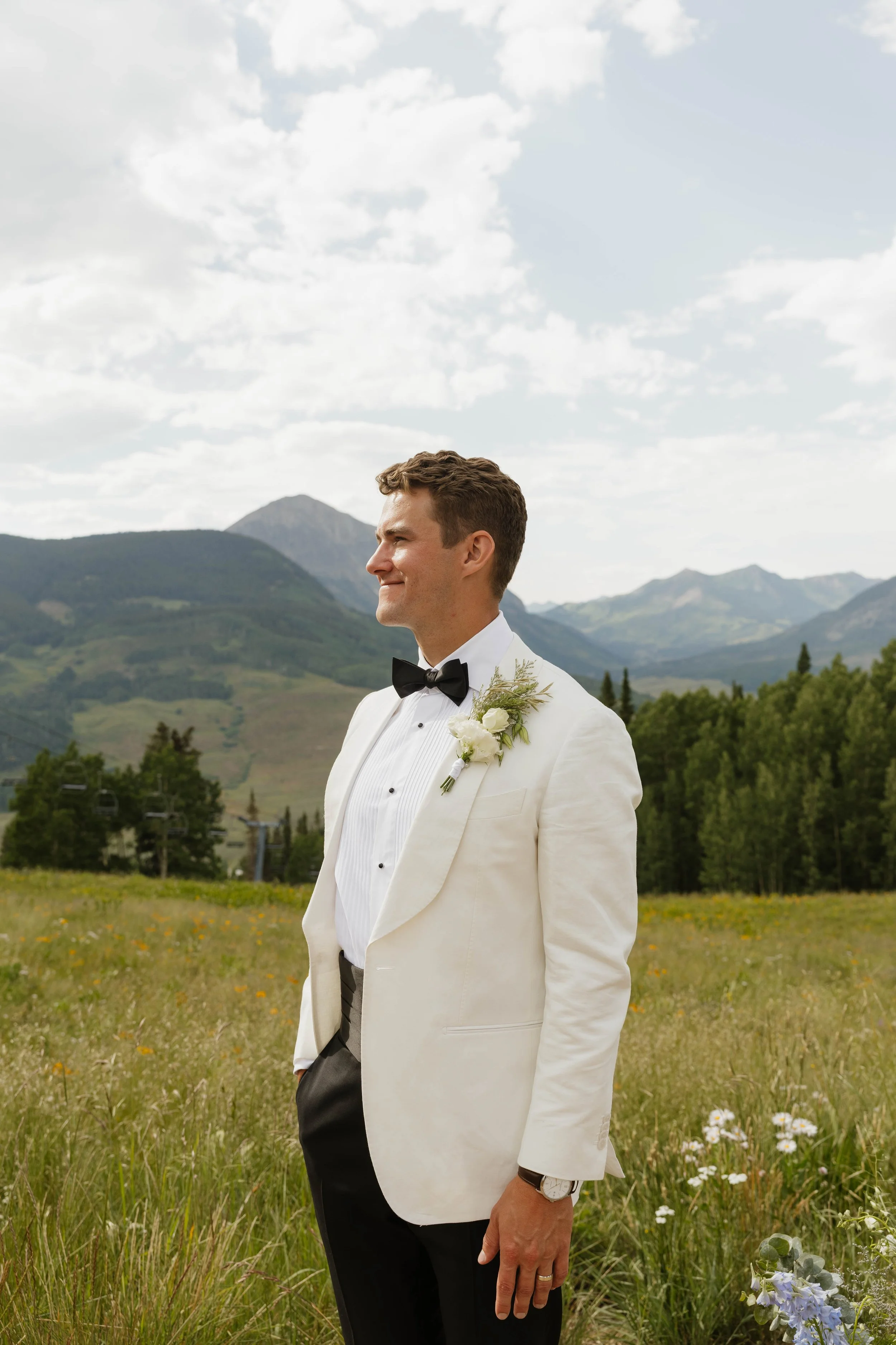A groom in a white tuxedo jacket, black bow tie, and black trousers stands outdoors in a grassy field with mountains and cloudy sky in the background.