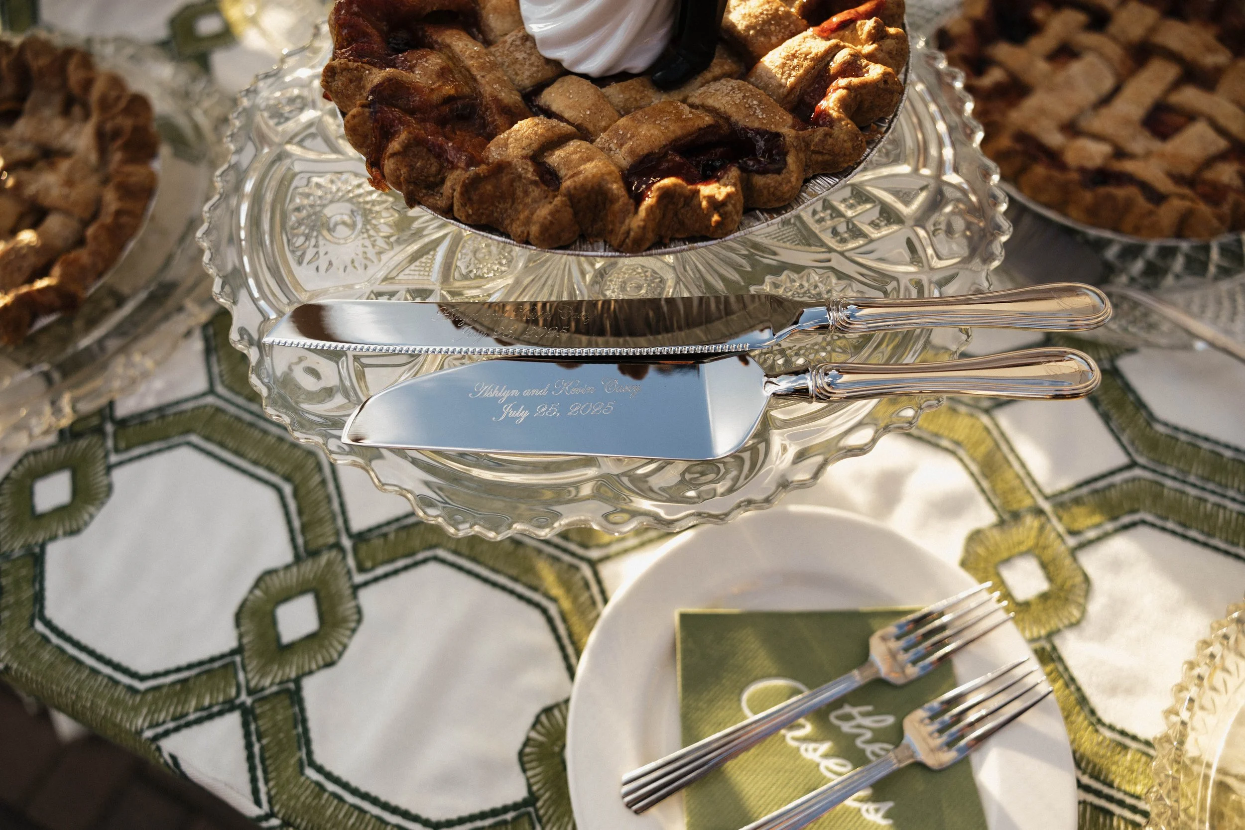 A glass cake stand with a lattice-topped cherry pie and a dollop of whipped cream on top, set on a table with a decorative tablecloth. Two serving utensils rest on a plate with two forks, and an engraved knife is placed on a nearby dish.