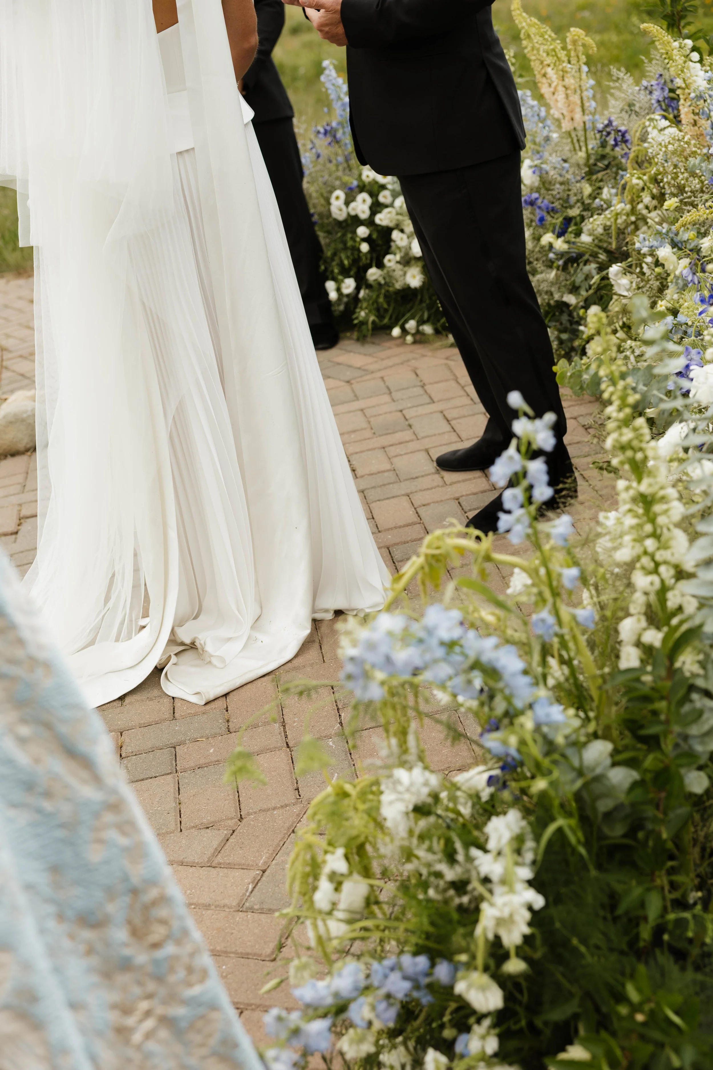 A bride in a white wedding dress and a groom in a black suit standing on a brick path surrounded by floral arrangements, during a wedding ceremony.