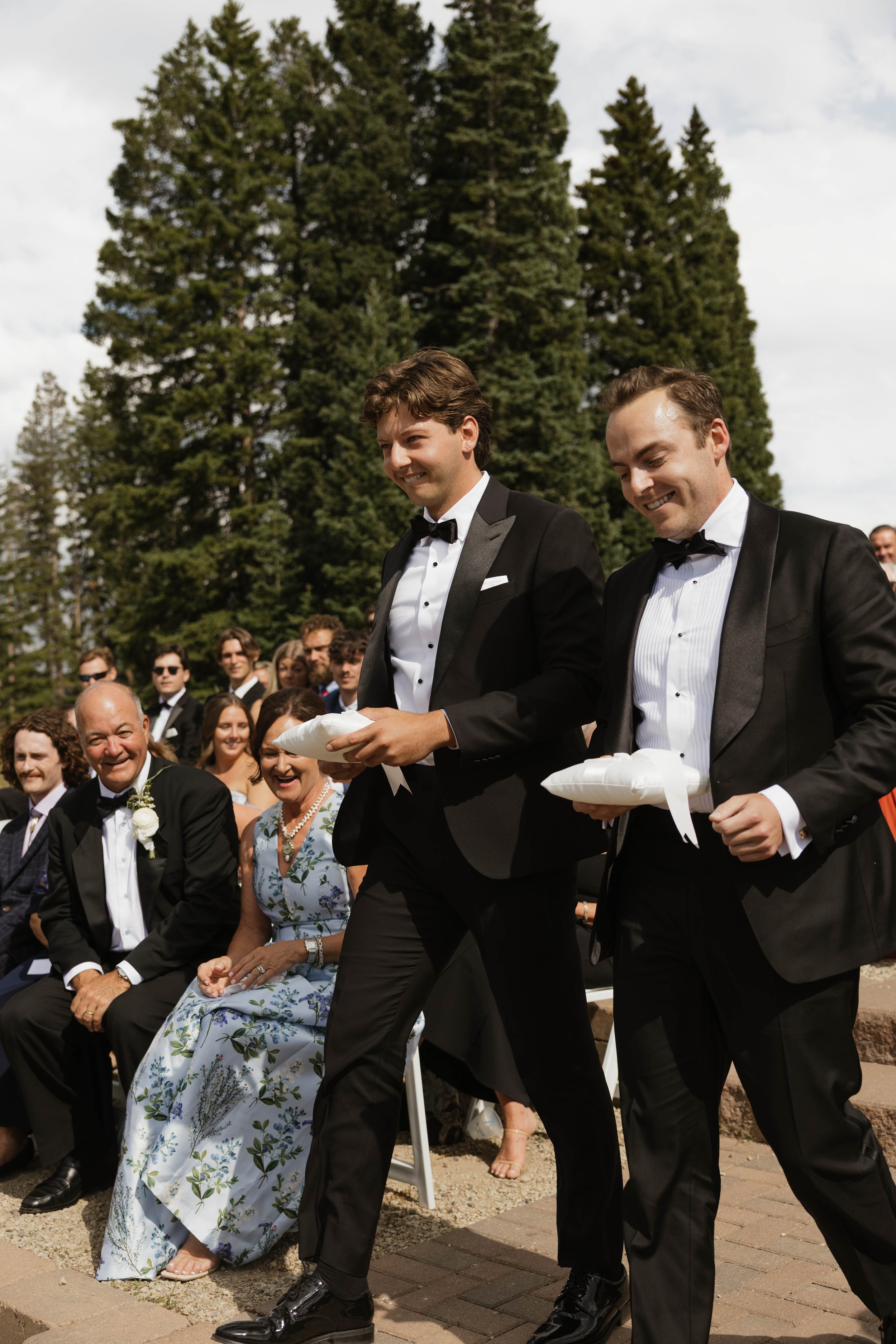 Two men in tuxedos walking in a wedding ceremony with guests seated in the background and tall trees