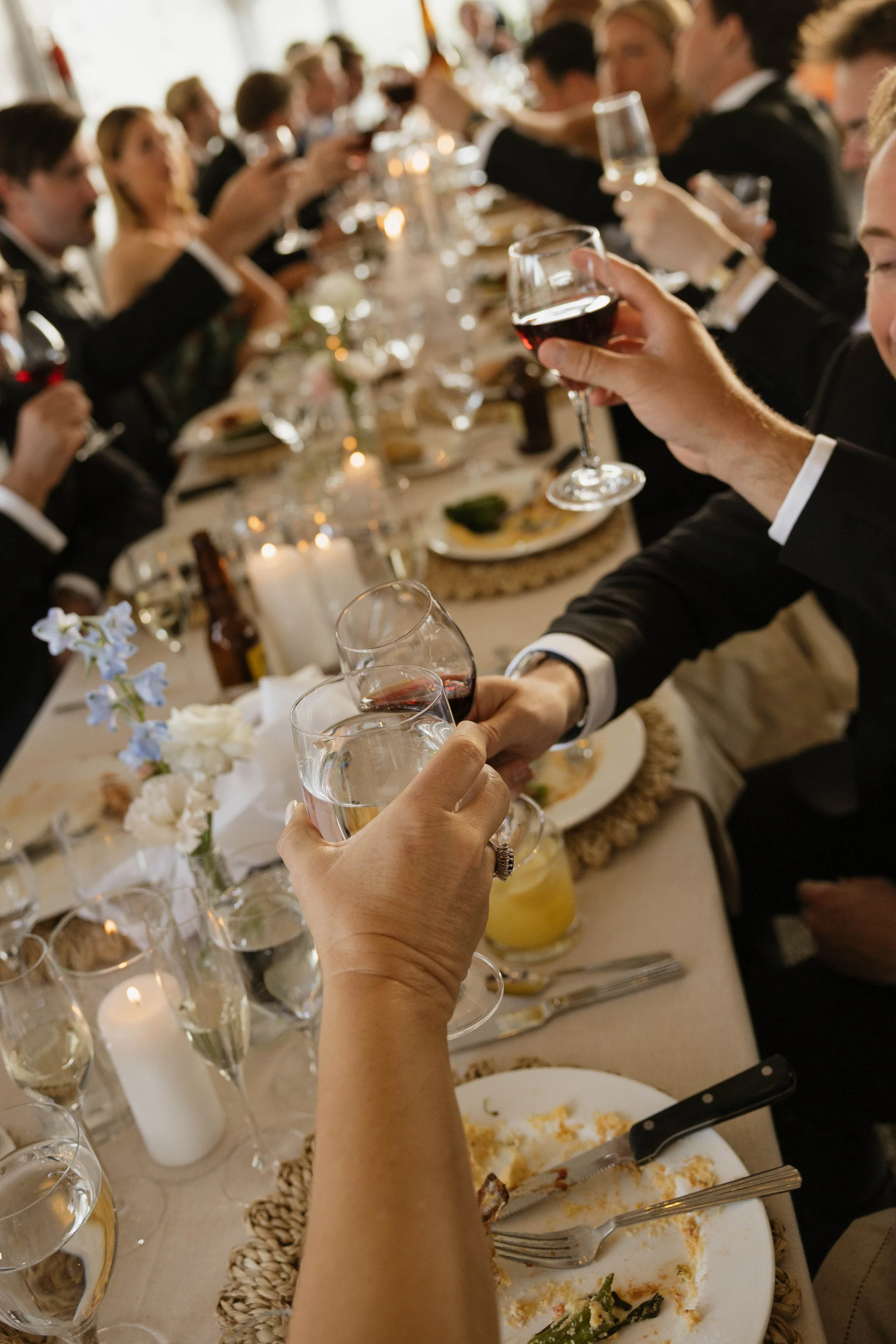 People at a formal dinner toasting with wine glasses, surrounded by candles, flowers, and partly eaten food on the table.