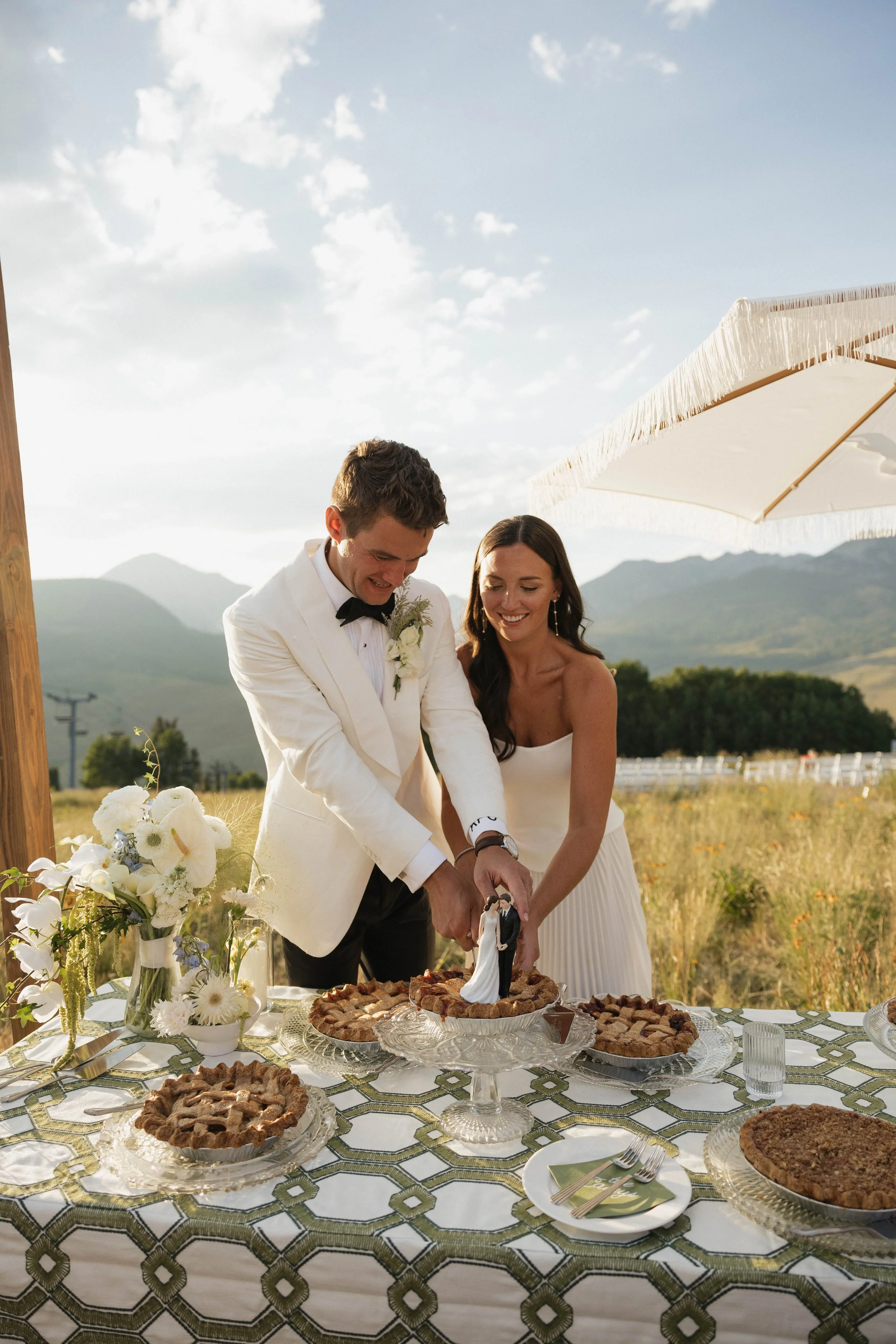 A newlywed couple is cutting their wedding cake outdoors in a scenic mountain setting, with white flowers on the table and a large umbrella providing shade.