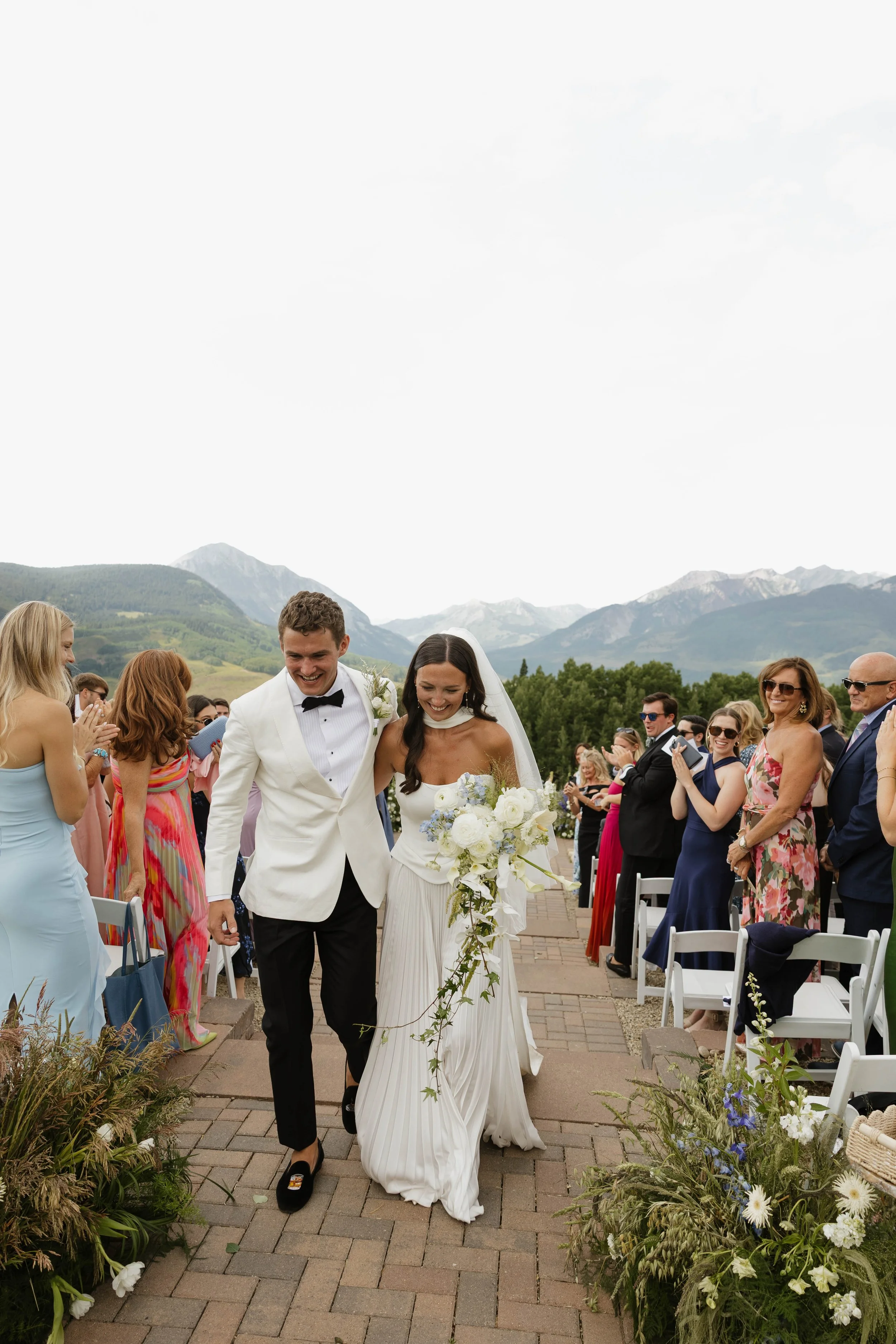 A bride and groom walking down the aisle surrounded by friends and family during an outdoor wedding ceremony, with scenic mountains and greenery in the background.