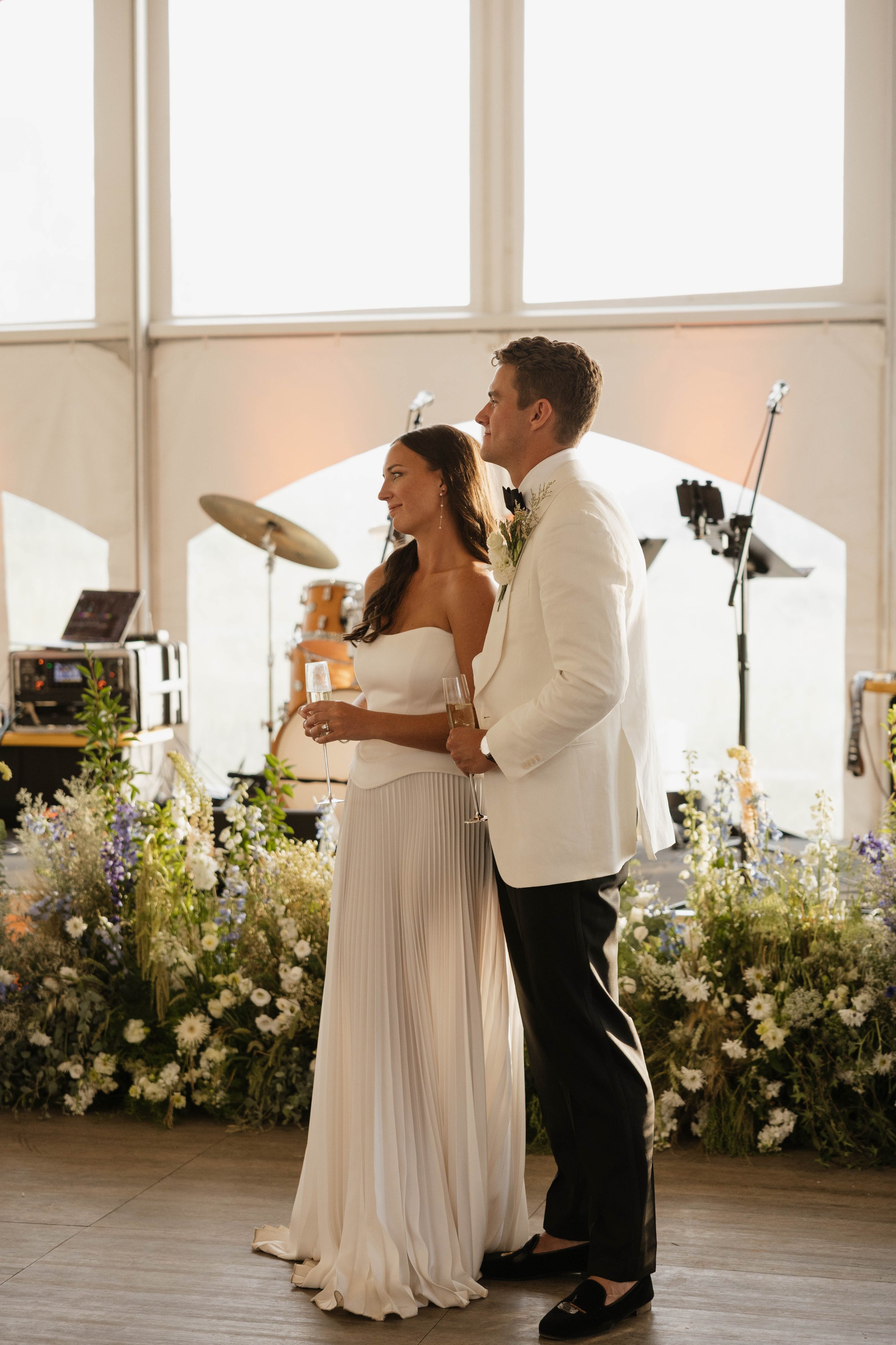 A bride and groom at their wedding reception holding champagne glasses, standing side by side in a decorated venue with musical instruments and flowers in the background.