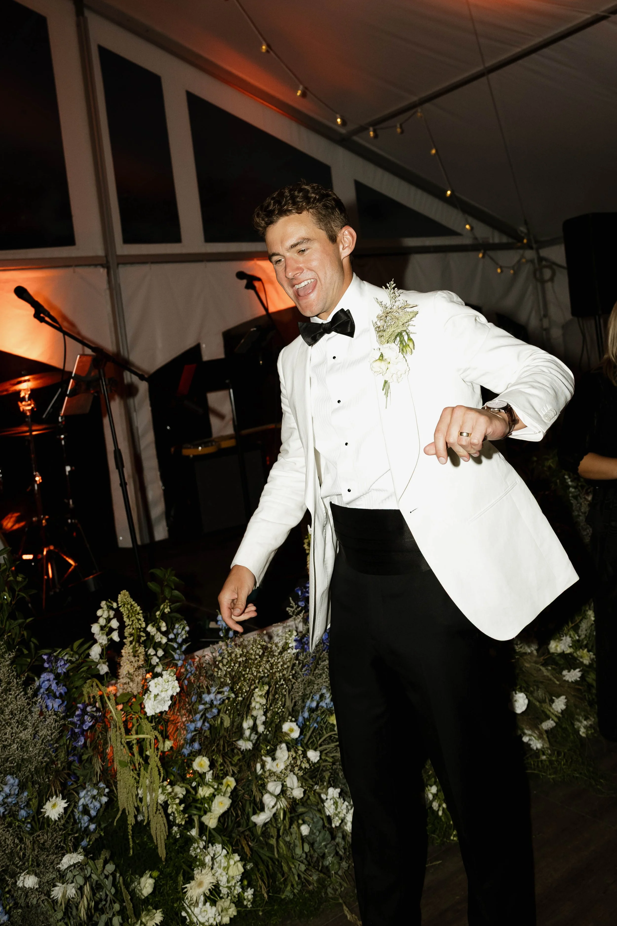 A man in a white tuxedo jacket, black bow tie, and black pants dancing at a formal event with a floral arrangement in front of him and a drum set and microphones in the background.